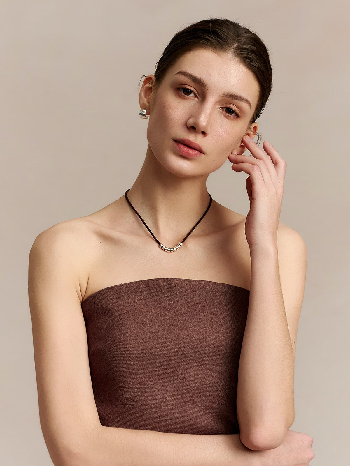 Model wearing Fairyokai Balance Glide leather and silver bead necklace, lifting her left hand to her ear and tilting her head slightly to the left, on a beige background.