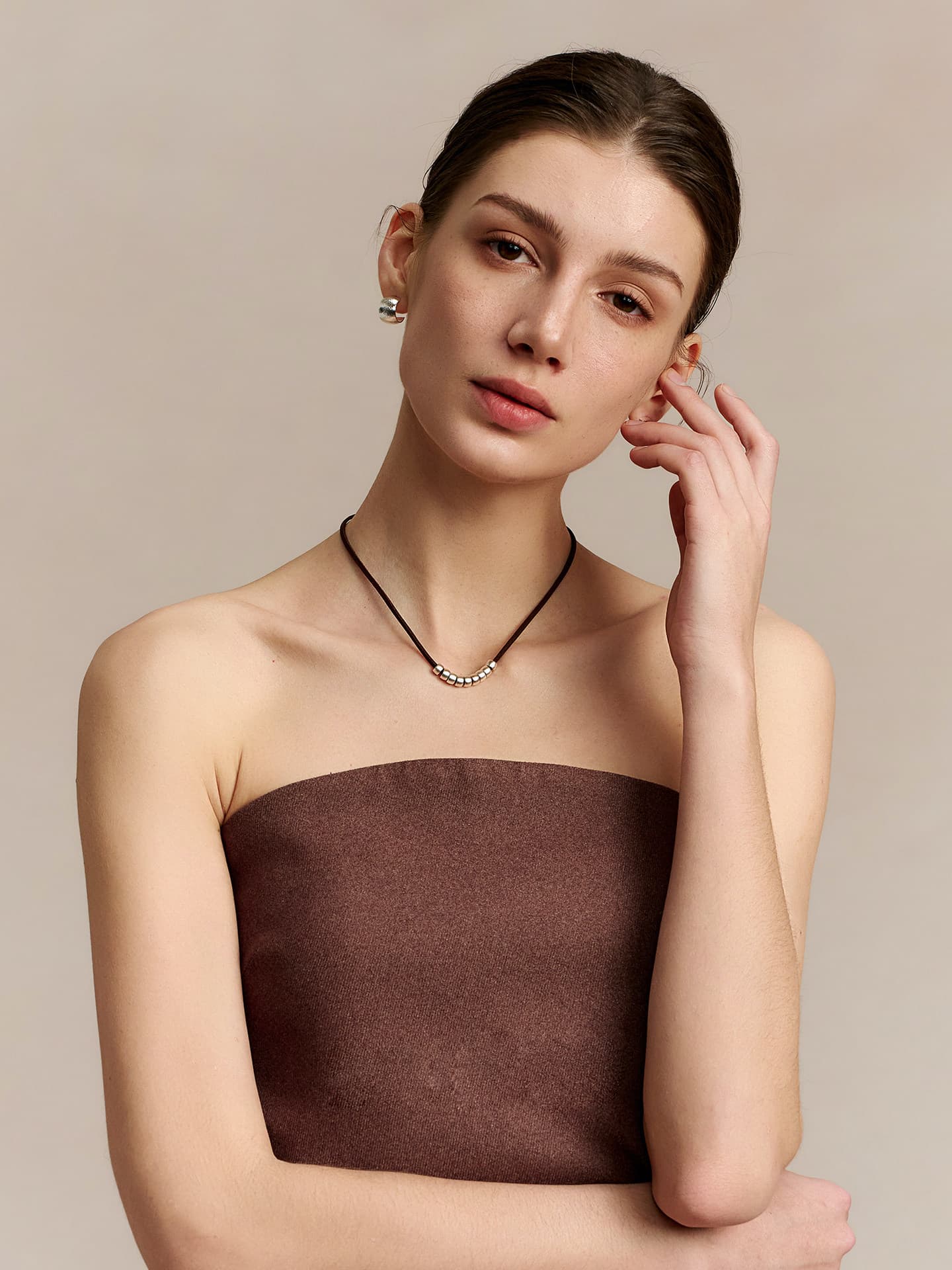 Model wearing Fairyokai Balance Glide leather and silver bead necklace, lifting her left hand to her ear and tilting her head slightly to the left, on a beige background.