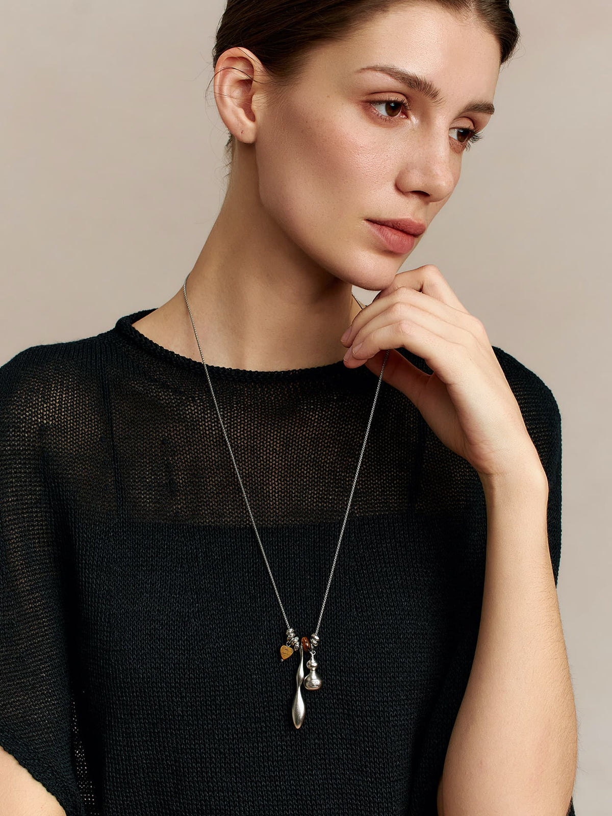Woman wearing Fairyokai Balance Veil tiger eye gourd necklace, resting her chin on her hand, looking slightly down to the left, dressed in a dark top, against a neutral background.
