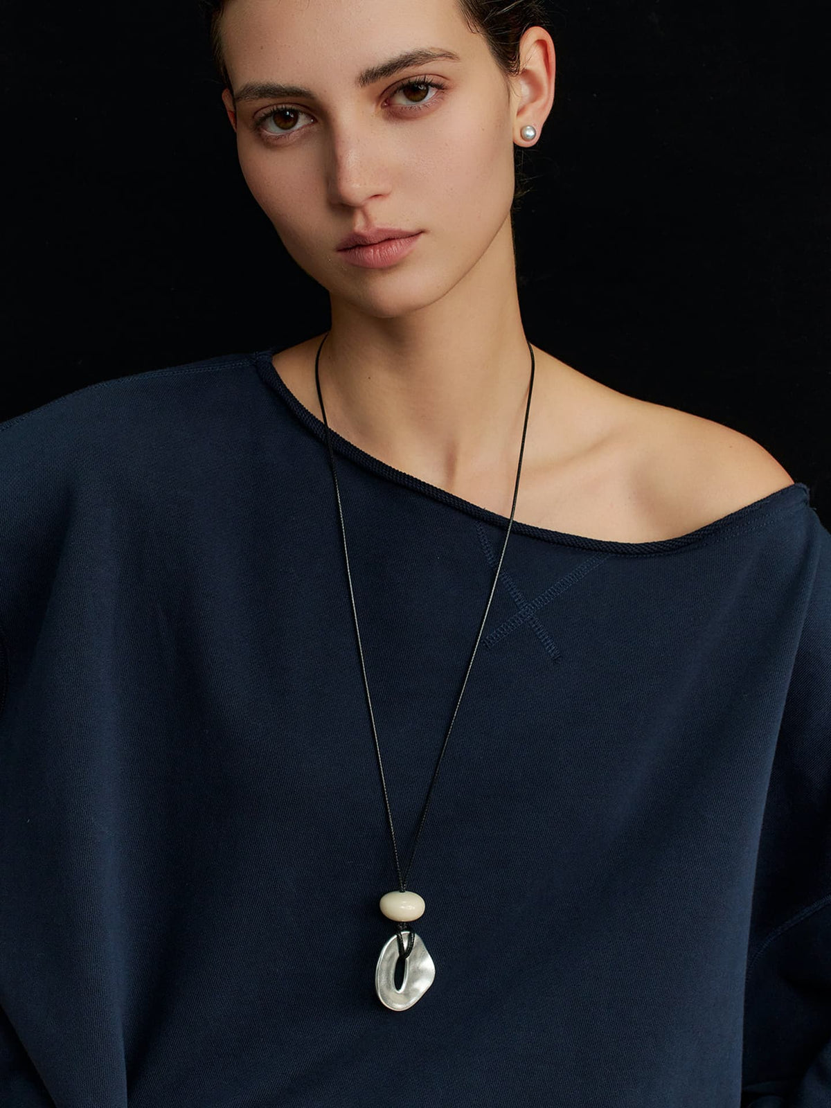 Woman wearing Fairyokai Breath Pendant adjustable waxed cord oval necklace, facing the camera in a dark top, left shoulder exposed, against a black background.