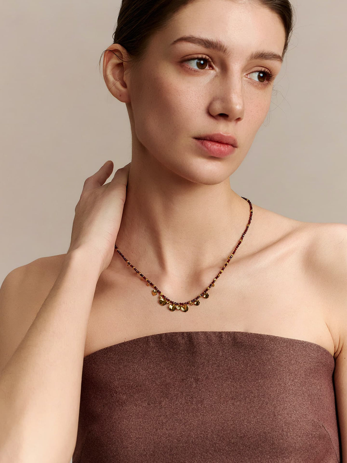 Model wearing Fairyokai Breeze Whisper gold leaf natural stone necklace, looking to the left with her right hand touching the back of her neck, dressed in a strapless top, on a neutral background.