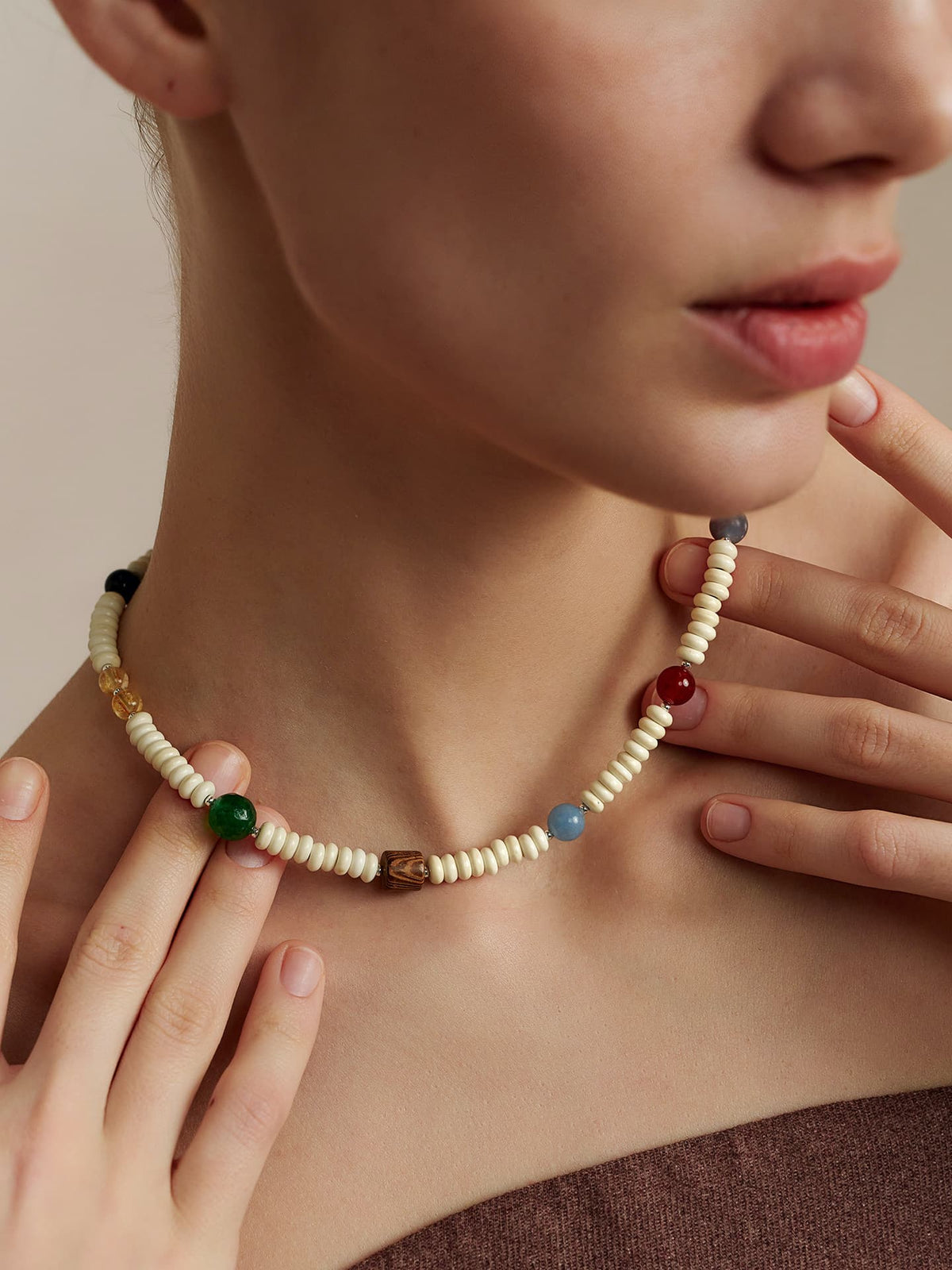 Close-up of a model's neck wearing the Fairyokai Color Rhythm multicolor stone roller bead necklace, dressed in a strapless top, with both hands touching the necklace, on a neutral background.
