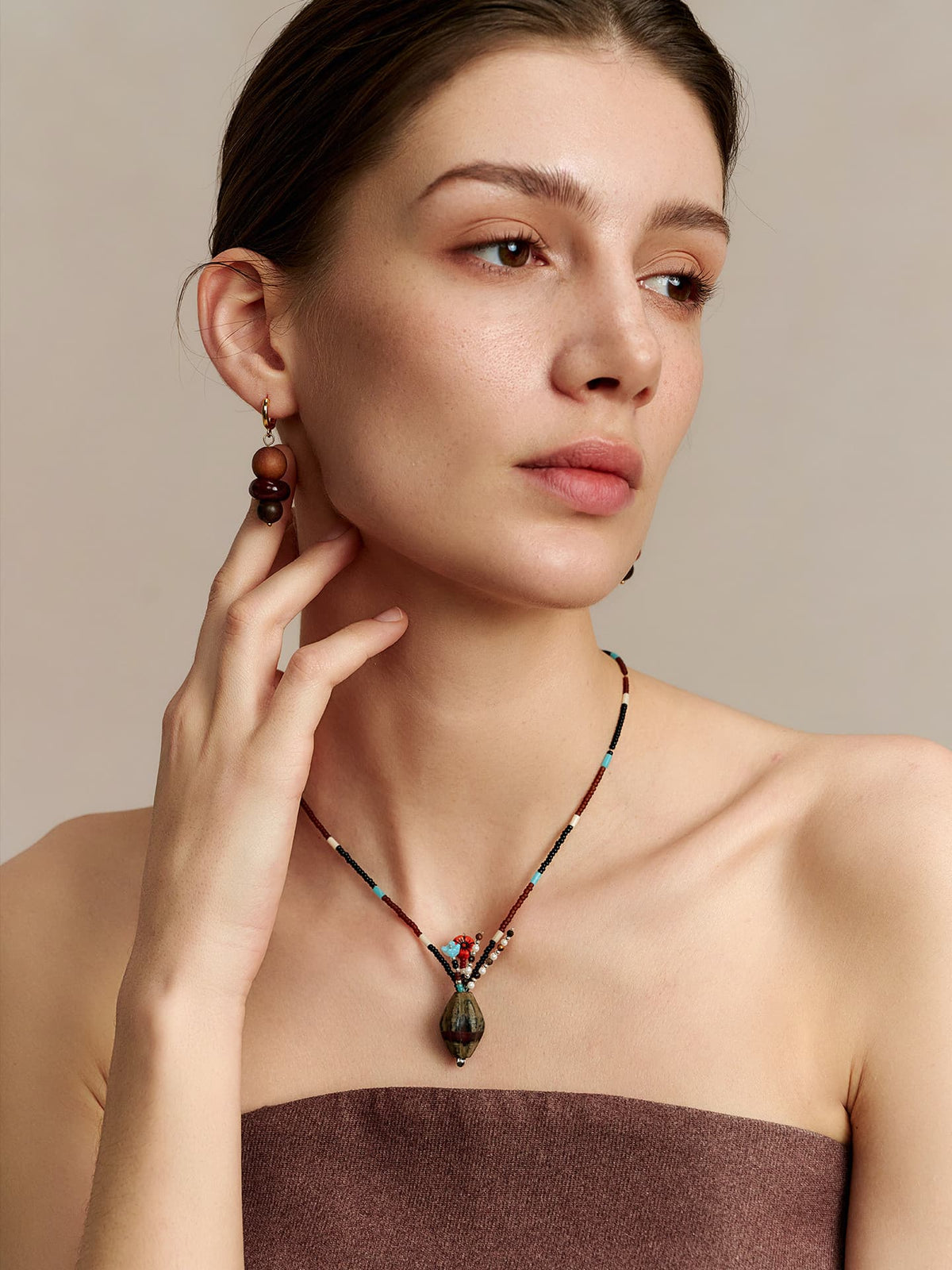 Model in a strapless top, looking to the left, touching her right ear, wearing the Fairyokai Culling the Cream agate vase pendant necklace, on neutral background.