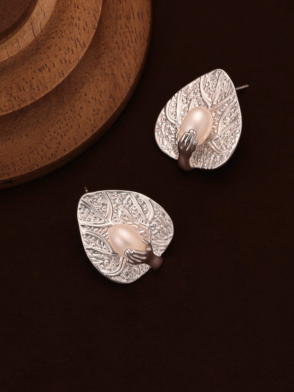 Top-down flat lay of Fairyokai Dew-Catching leaf and palm earrings on a deep brown background, with a wooden display stand partially visible in the upper left corner.