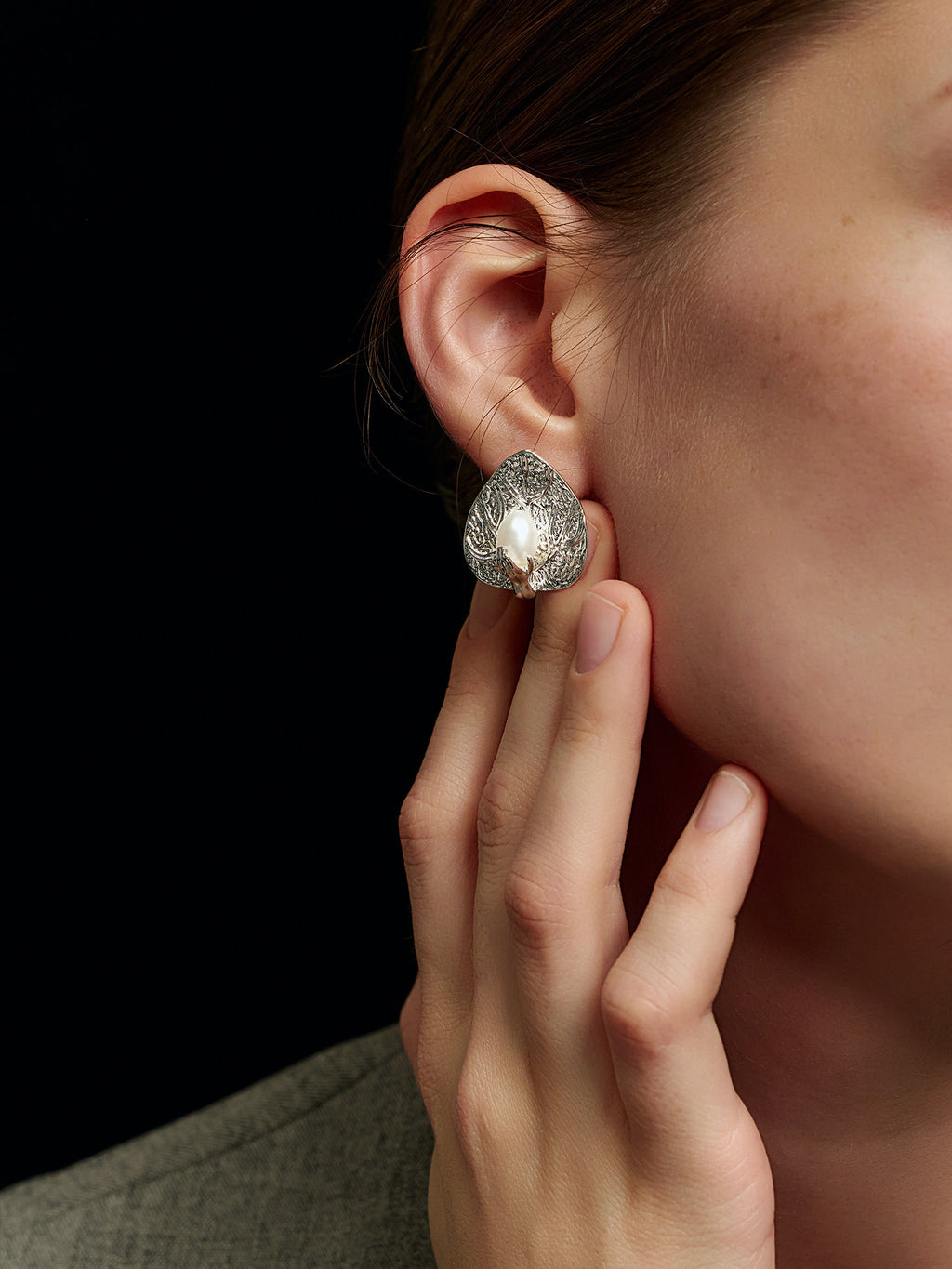 Close-up of a model's right hand touching her right ear, wearing Fairyokai Dew-Catching leaf and palm earrings, on a black background.