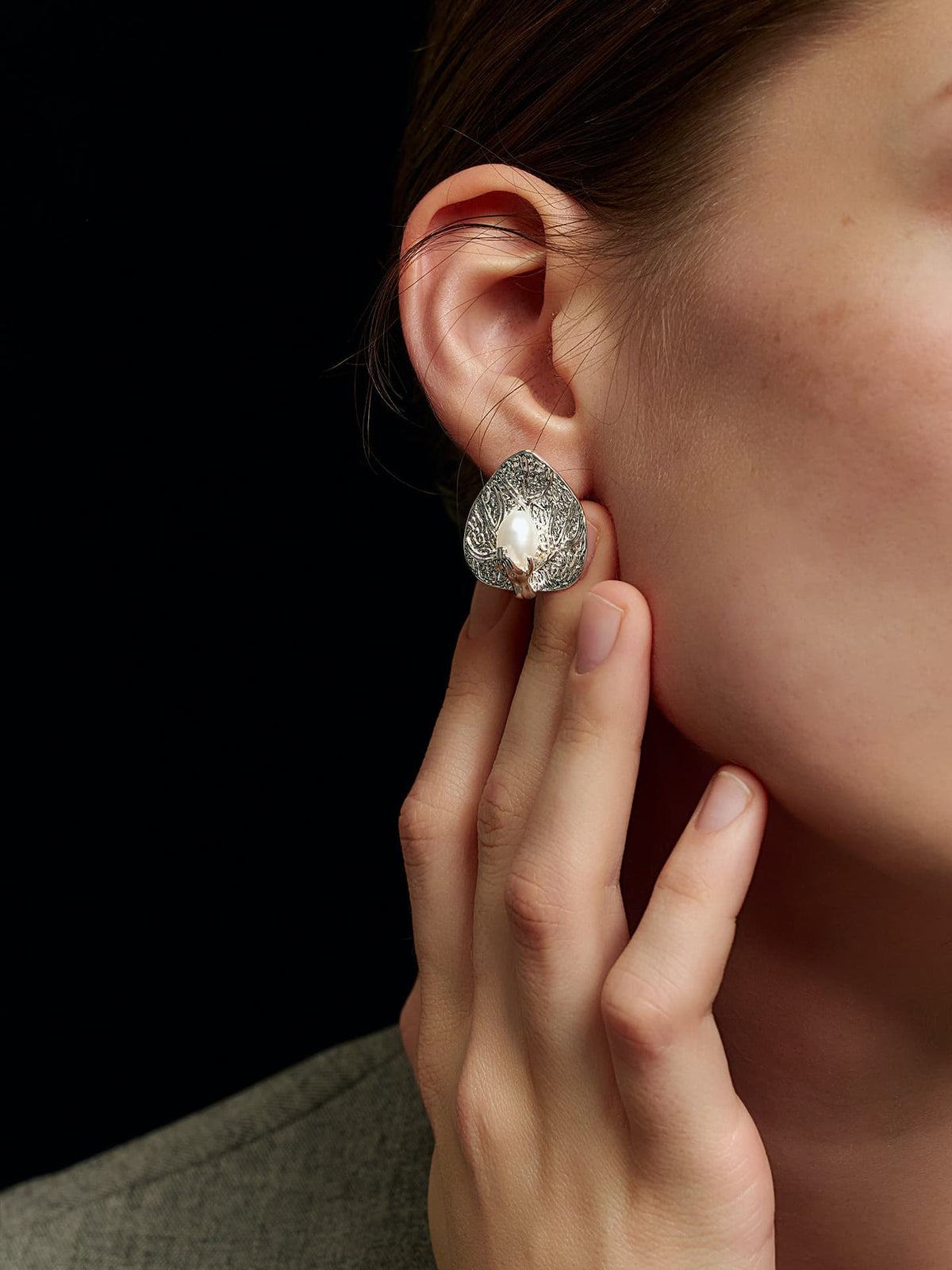 Close-up of a model's right hand touching her right ear, wearing Fairyokai Dew-Catching leaf and palm earrings, on a black background.