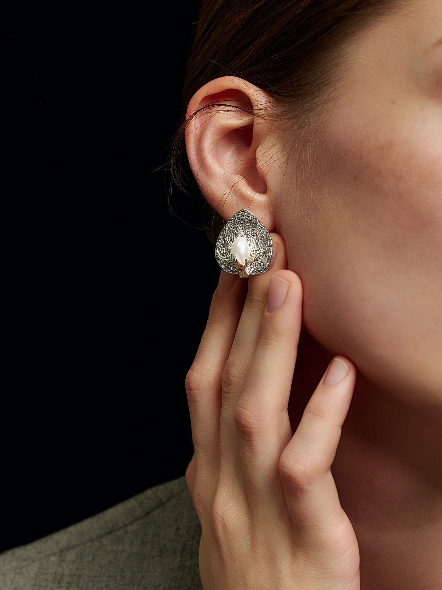 Close-up of a model's right hand touching her right ear, wearing Fairyokai Dew-Catching leaf and palm earrings, on a black background.