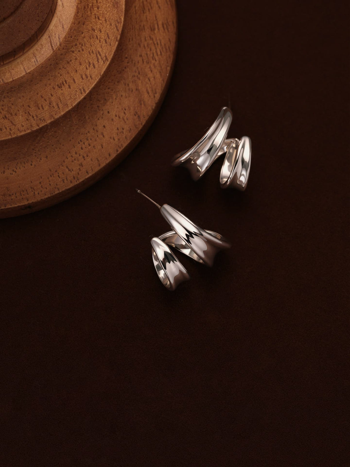 Fairyokai Double Arc sculptural open C-shape hoop earrings flat-laid on a deep brown background, with a wooden display stand partially visible in the upper left corner.