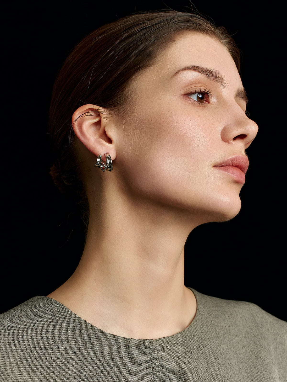 Close-up of a model in a solid sleeveless top wearing Fairyokai Double Arc C-shape hoop earrings, looking up and to the left, with focus on her right profile against a black background.