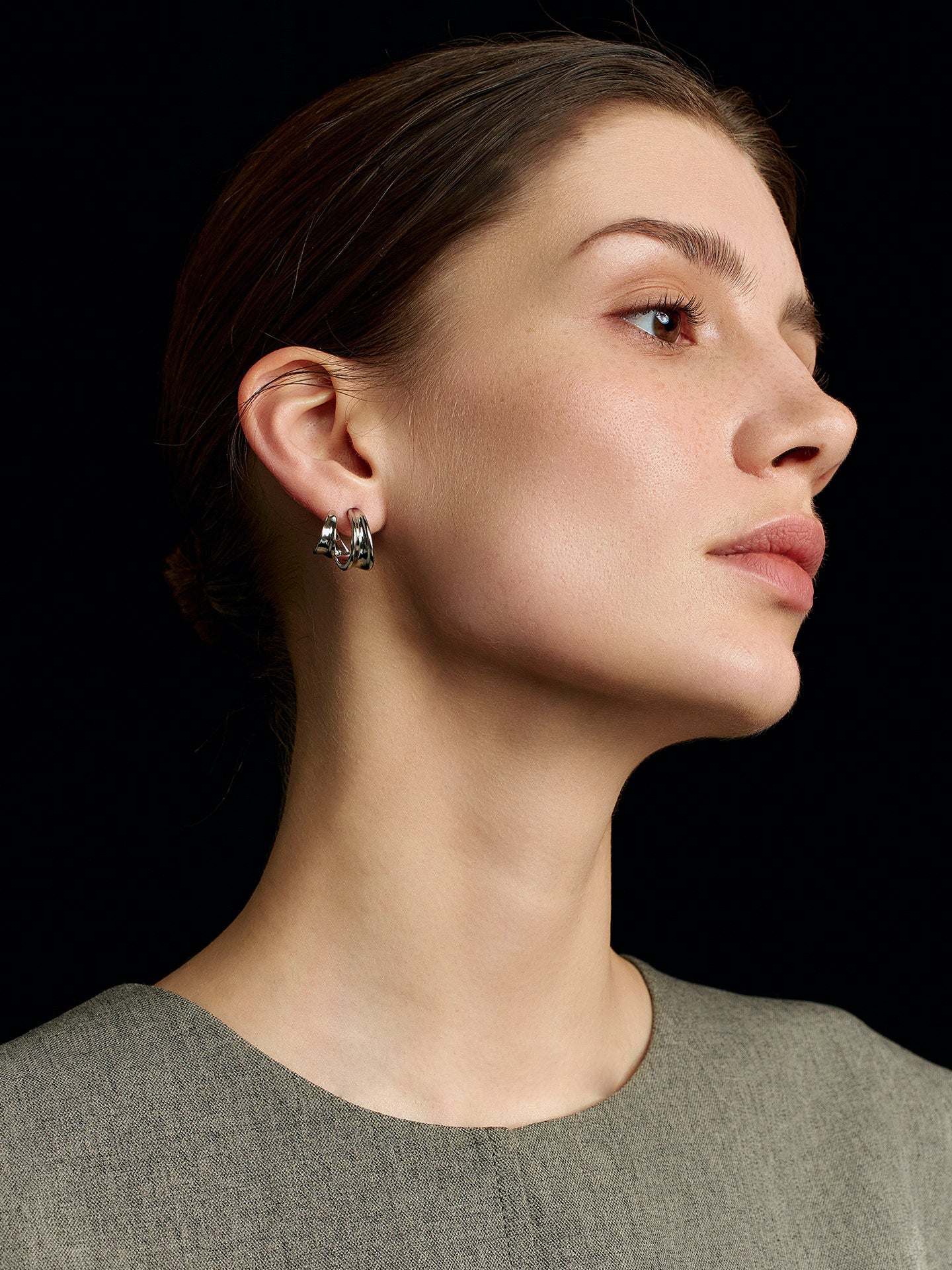 Close-up of a model in a solid sleeveless top wearing Fairyokai Double Arc C-shape hoop earrings, looking up and to the left, with focus on her right profile against a black background.