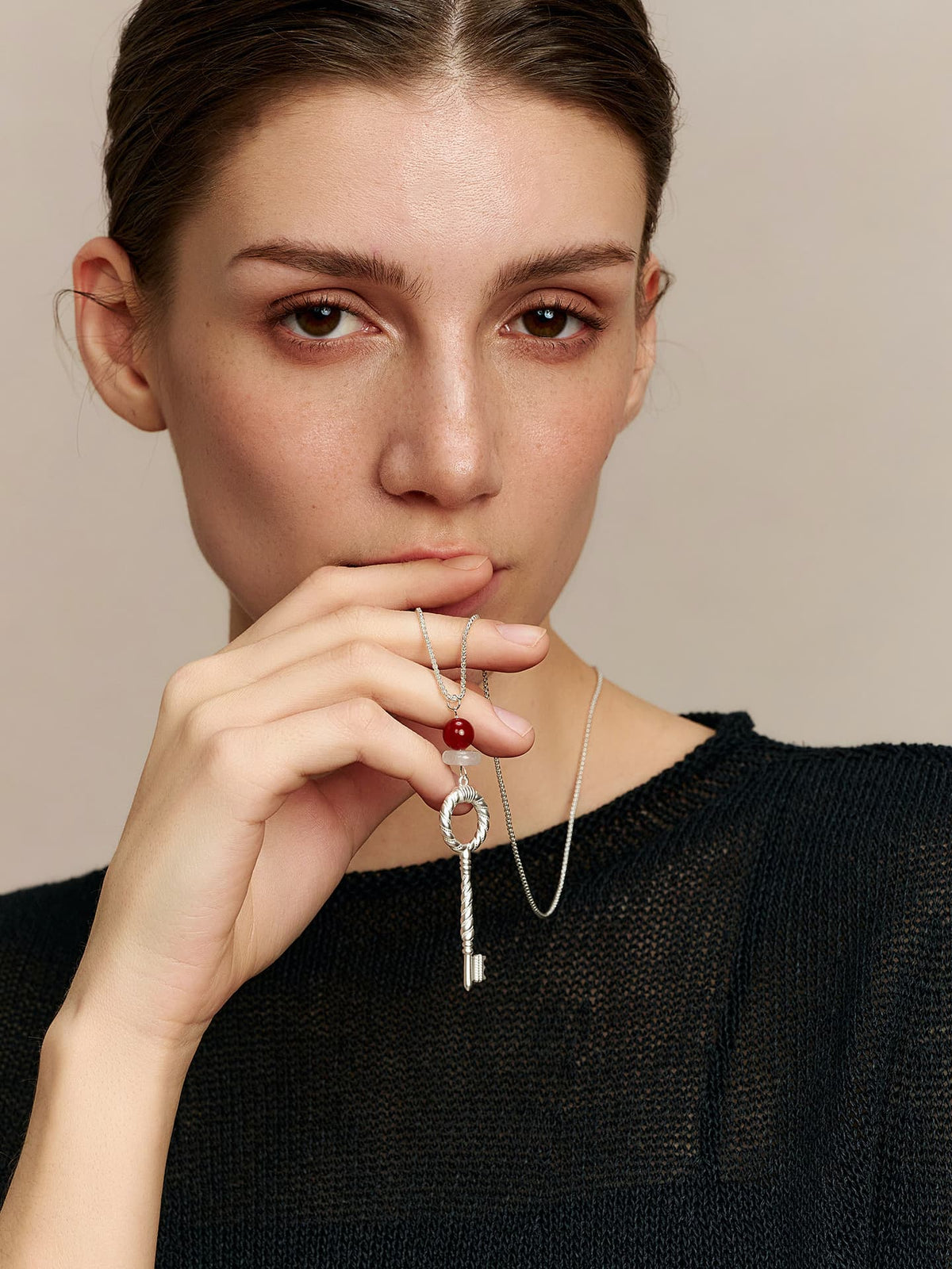 Model in a black top, wearing the Fairyokai Dream Key red agate crystal key pendant necklace, right hand slightly raised holding the pendant near her mouth, facing the camera, on neutral background.