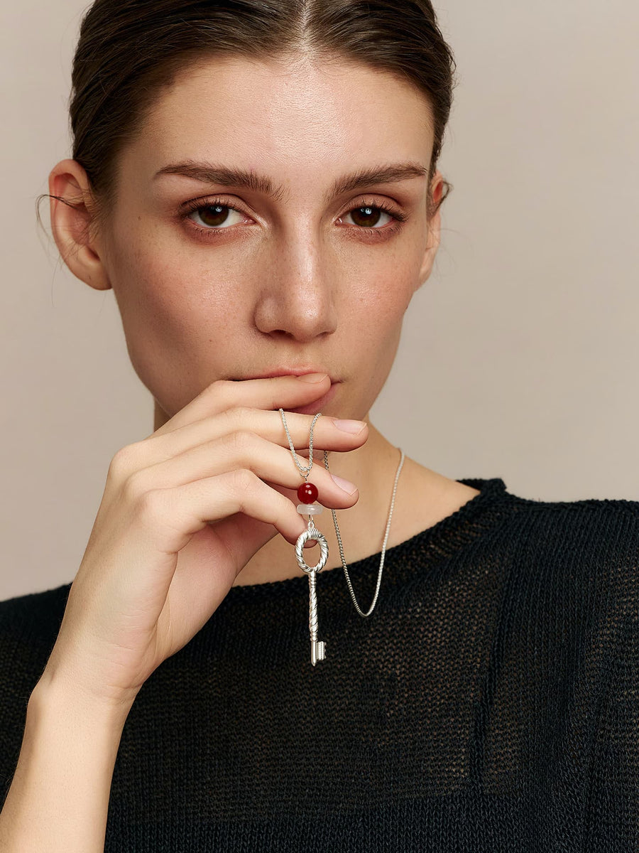 Model in a black top, wearing the Fairyokai Dream Key red agate crystal key pendant necklace, right hand slightly raised holding the pendant near her mouth, facing the camera, on neutral background.