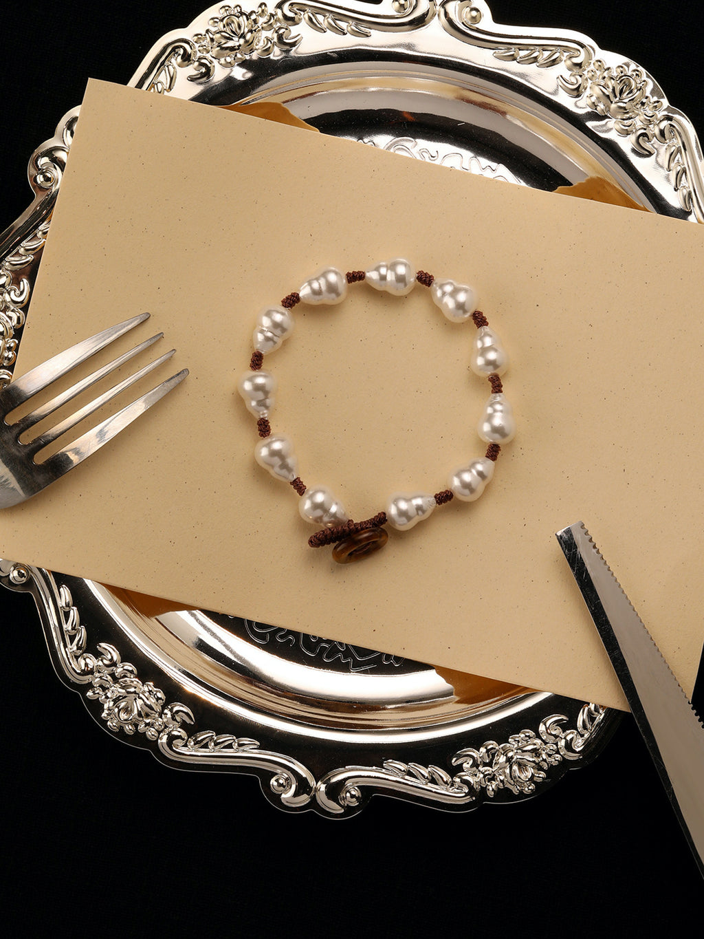 Fairyokai Full Embrace faux pearl bracelet flat-laid on a beige card with a silver tray and tableware, showing the gourd-shaped pearl beads.