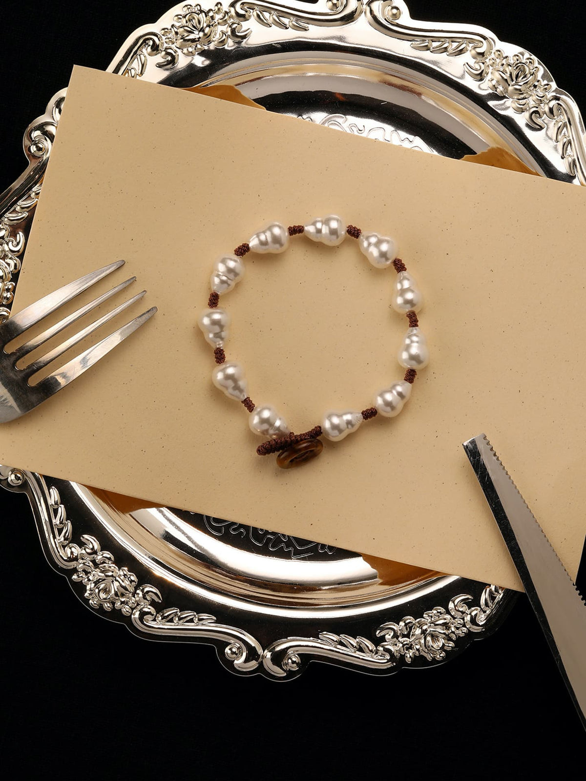 Fairyokai Full Embrace faux pearl bracelet flat-laid on a beige card with a silver tray and tableware, showing the gourd-shaped pearl beads.