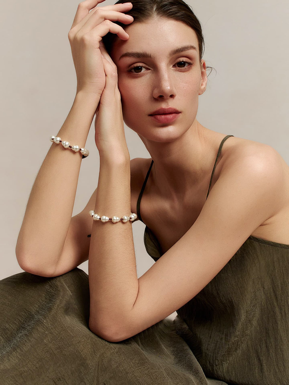 Model in an olive dress wearing Fairyokai Full Embrace faux pearl bracelets on both wrists, seated with arms crossed softly.