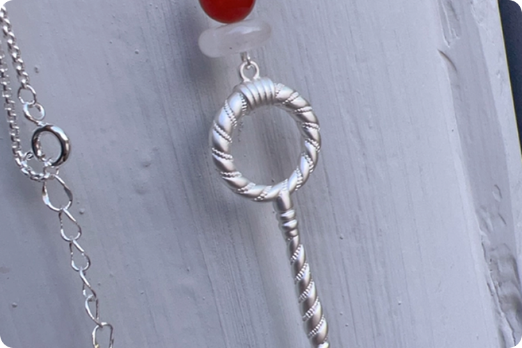 Close-up of a hanging Fairyokai Red Agate Crystal Key Pendant Necklace, on white wall background.