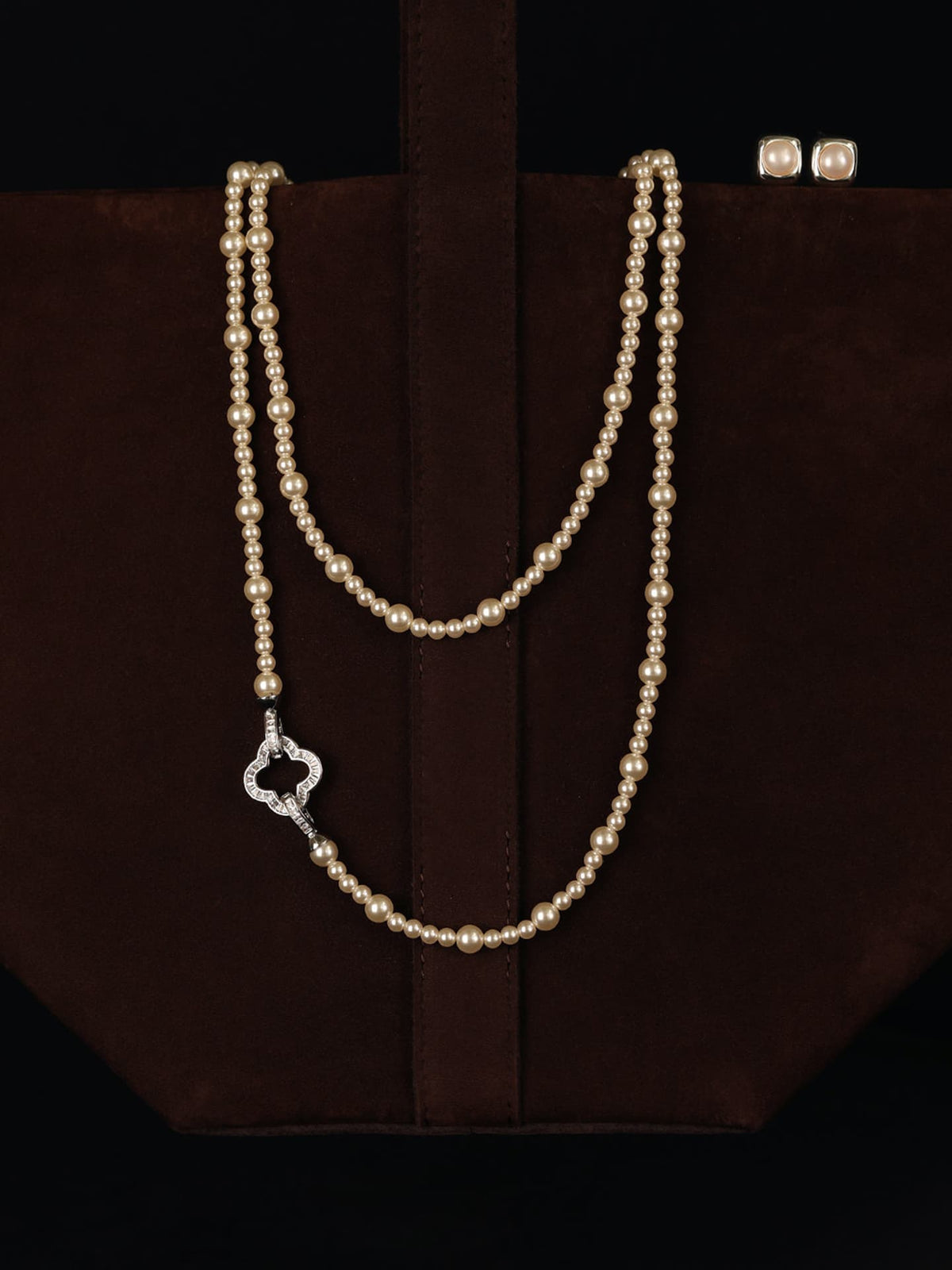 Fairyokai Layered Abundance alternating faux pearl necklace draped over a dark brown bag against a black background.