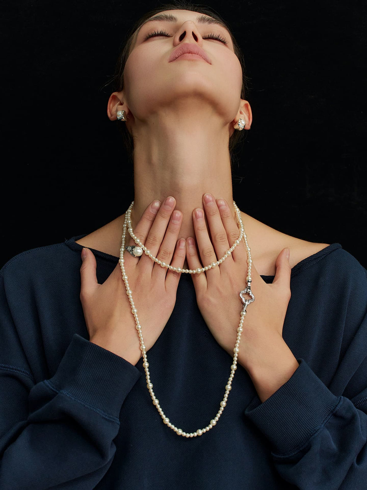 Model in a navy top, eyes closed and head tilted upward, touching the Fairyokai Layered Abundance alternating faux pearl necklace with both hands, immersed against a black background.