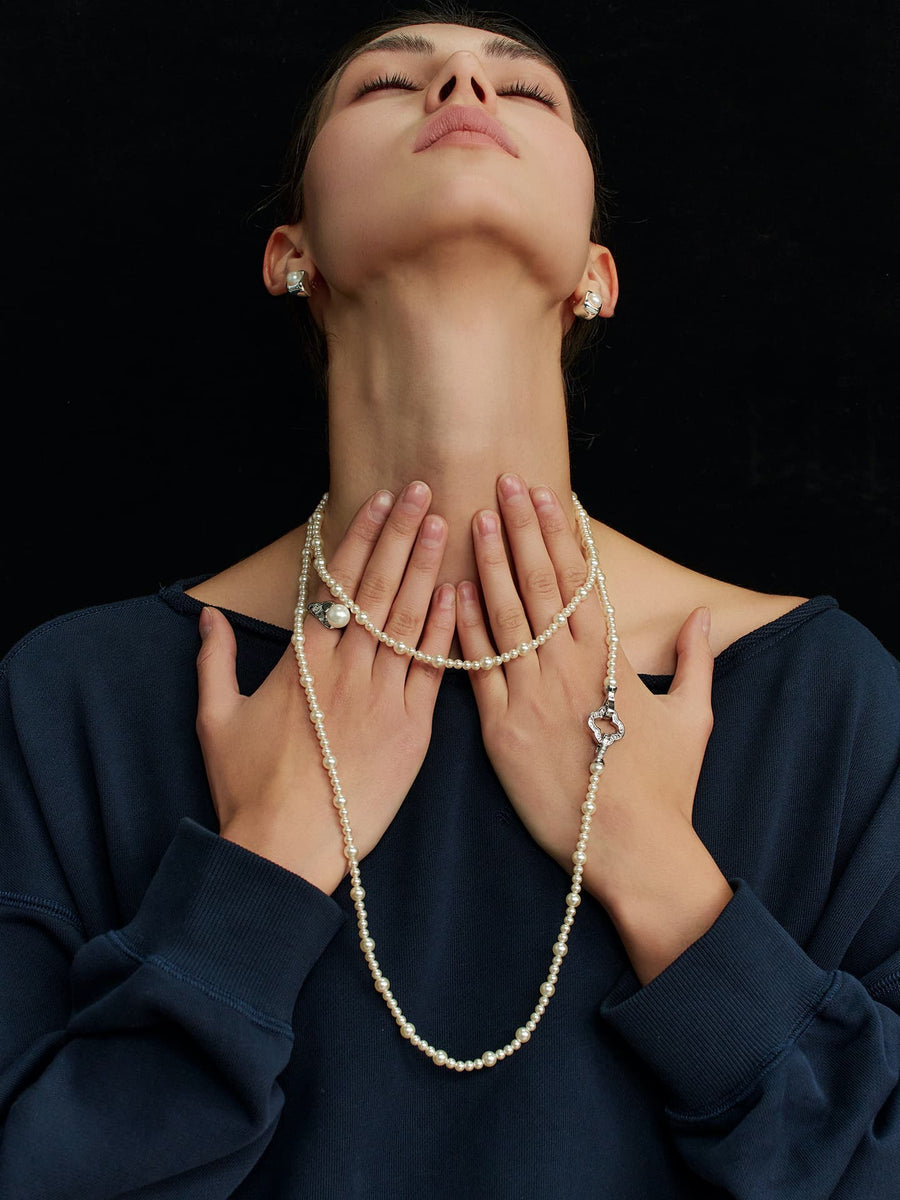 Model in a navy top, eyes closed and head tilted upward, touching the Fairyokai Layered Abundance alternating faux pearl necklace with both hands, immersed against a black background.