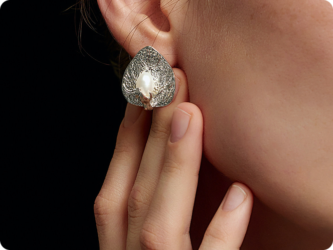 Close up of a woman wearing with Fairyokai dew catching leaf palm earrings, her fingers touching the earring, on a black background.