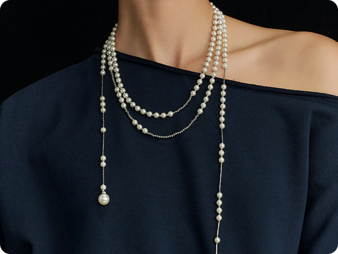 Close up of a woman wearing Fairyokai layered breath adjustable double -ayer faux pearl necklace, draped over a dark blue off-shoulder top, on a dark background