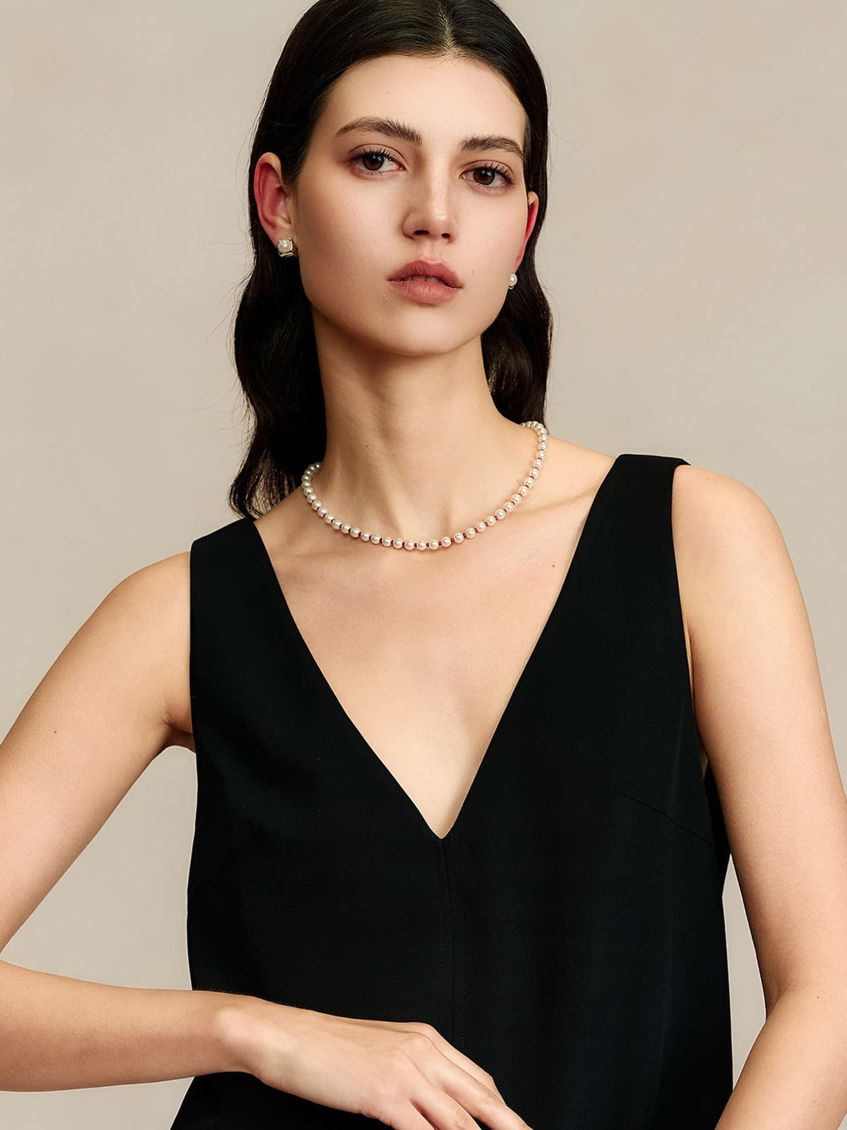 Model in a black V-neck sleeveless top wearing the Fairyokai Lunar Chord red Czech bead and jade pearl necklace, standing and facing the camera with her right hand slightly raised holding a black bag, framed from abdomen up.