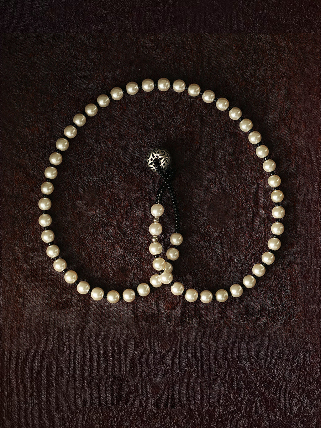 Fairyokai Lunar Chord double-layer faux pearl bracelet flat-laid on a dark brown textured background, showing the black cord and silver charm detail.