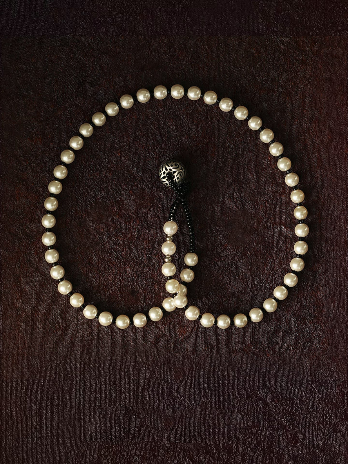 Fairyokai Lunar Chord double-layer faux pearl bracelet flat-laid on a dark brown textured background, showing the black cord and silver charm detail.