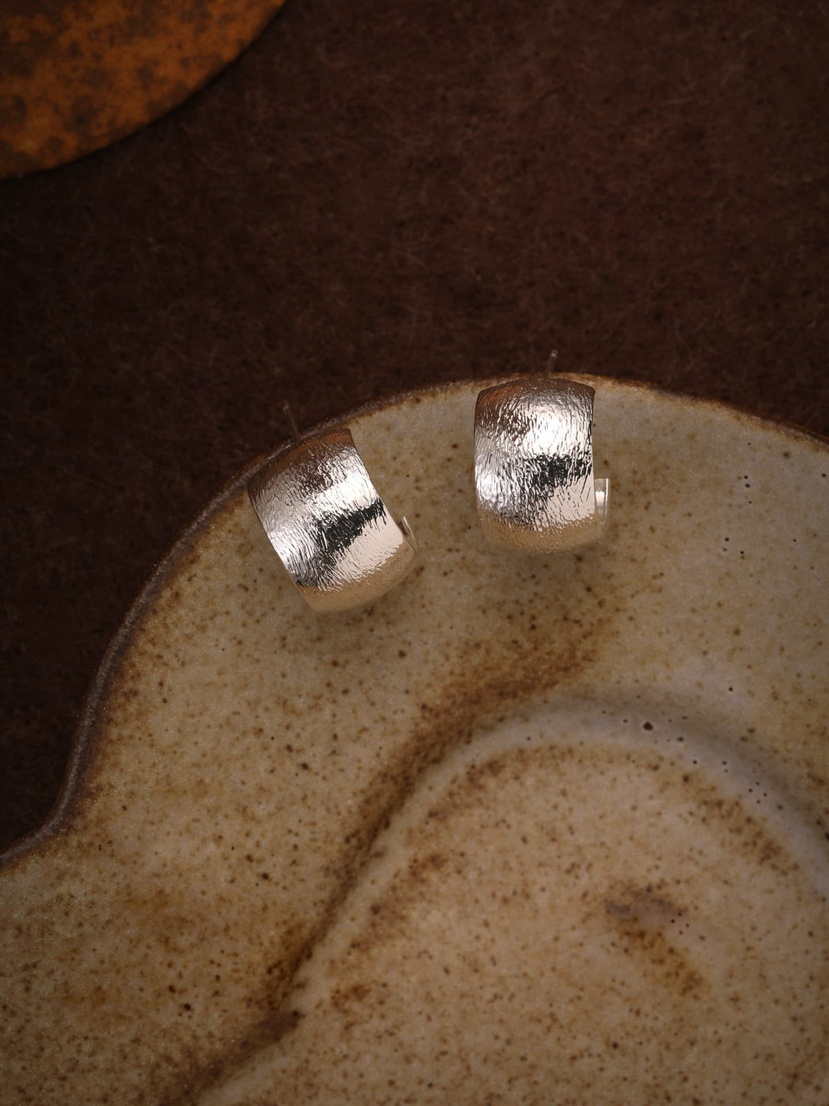 Fairyokai Moon Trace sand-textured C-shape hoop earrings flat-laid on a brown ceramic plate against a dark brown background, top-down view.