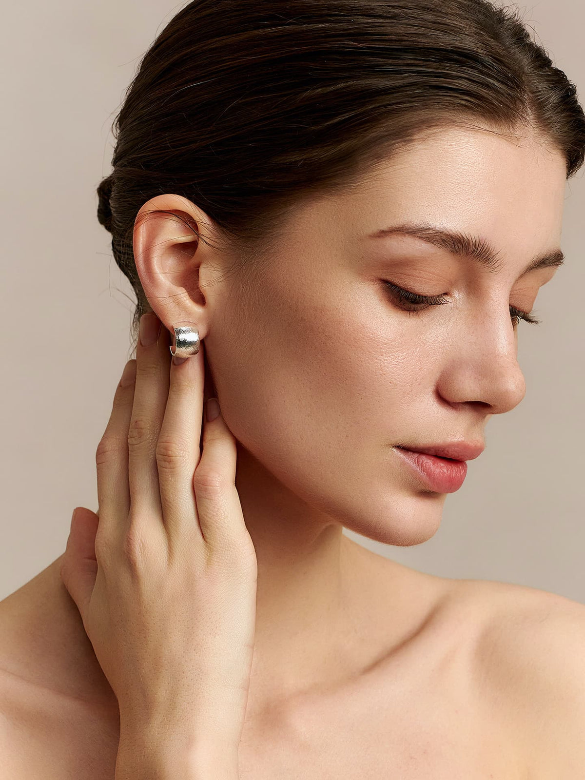 Model in a strapless top wearing Fairyokai Moon Trace sand-textured C-shape hoop earrings, looking down to the left with her right hand touching her right ear, side view on a neutral background