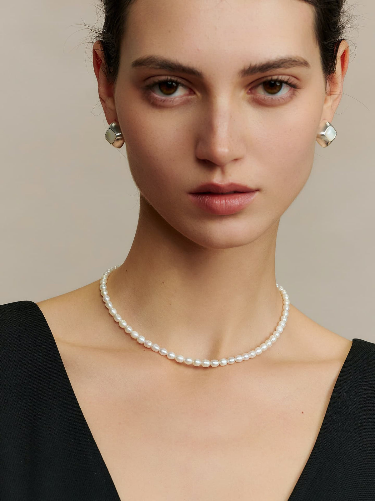 Model in a deep V-neck black dress, wearing Fairyokai Mother-of-Pearl iridescent stud earrings and a cream-colored pearl necklace, facing the camera, front view, against a neutral background