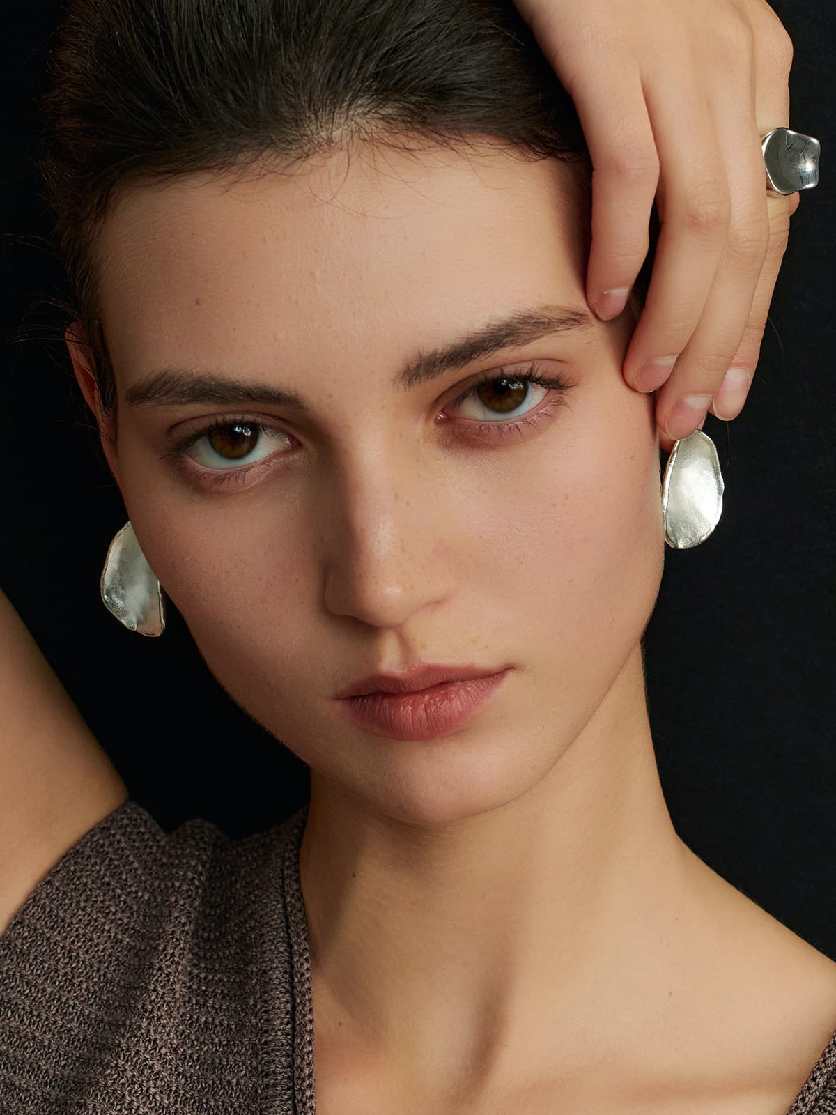 Close-up of a model facing the camera, right hand touching her left ear, wearing Fairyokai Pillow Stone matte slice earrings, framed from neck up on a black background.