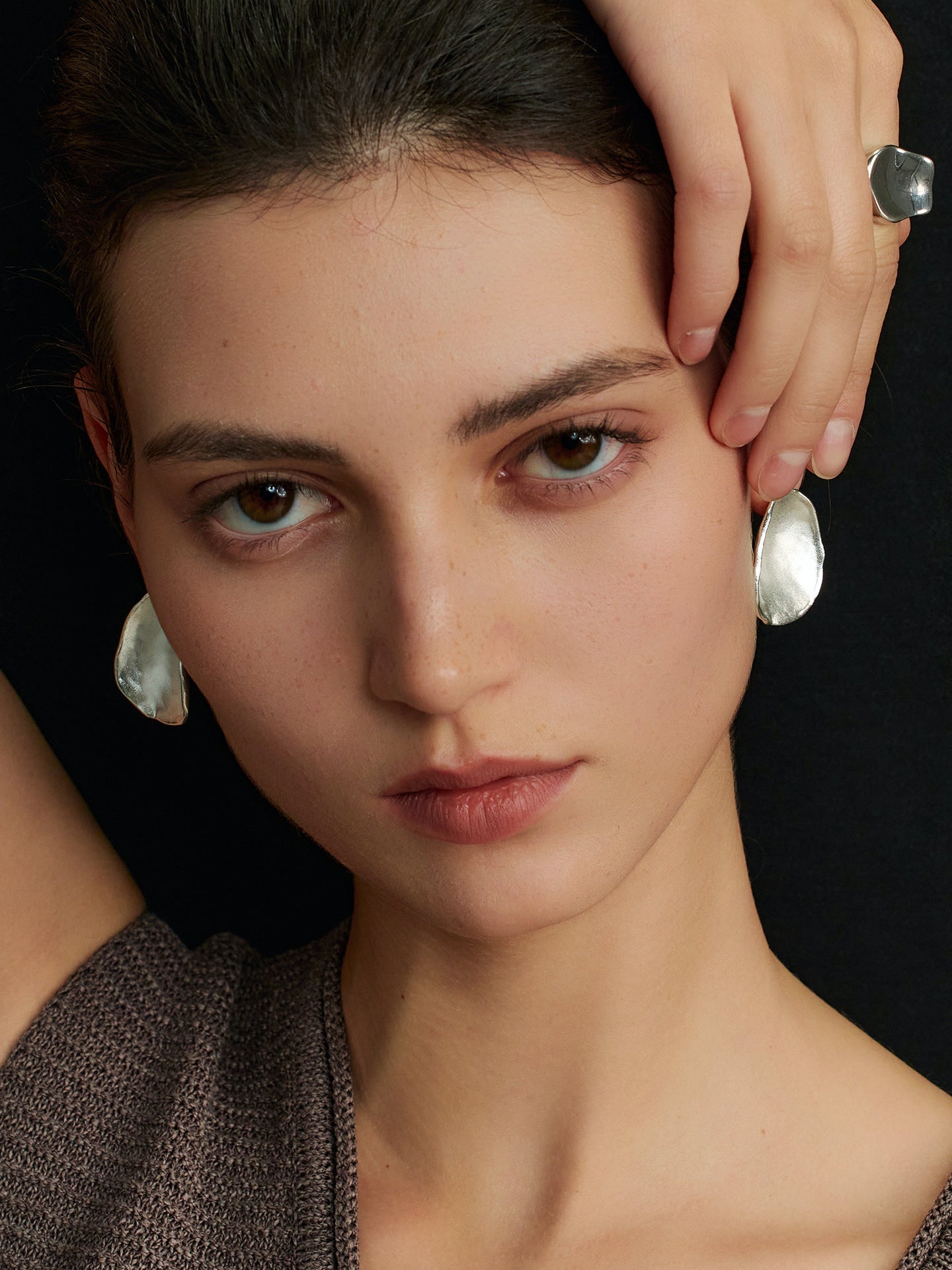 Close-up of a model facing the camera, right hand touching her left ear, wearing Fairyokai Pillow Stone matte slice earrings, framed from neck up on a black background.