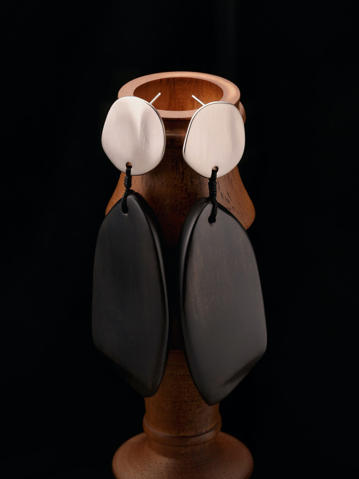 Fairyokai Quiet Leaf silver disc and ebony leaf earrings hanging on a wooden display stand against a black background.