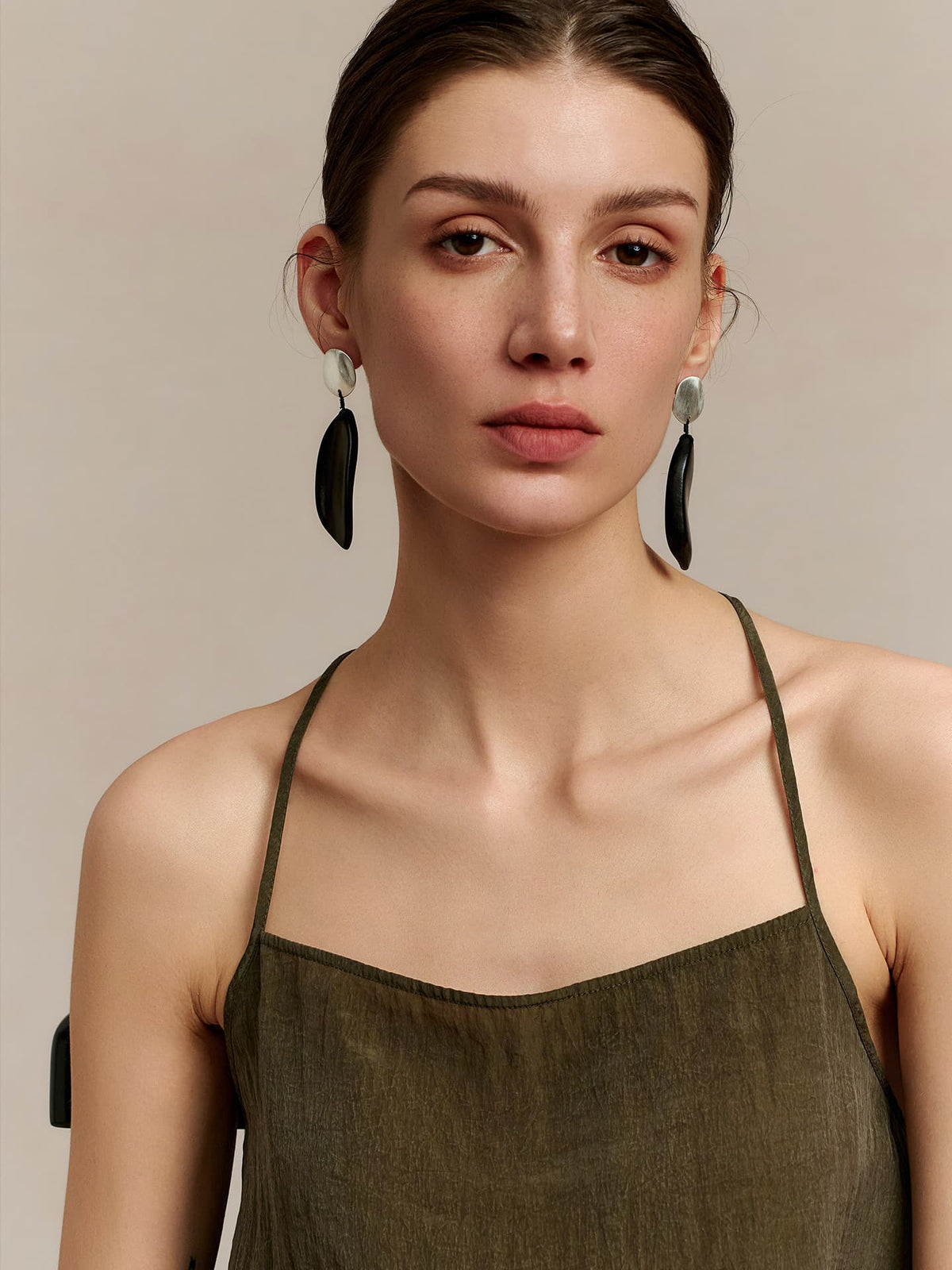 Model in a deep green thin-strap dress wearing Fairyokai Quiet Leaf silver disc and ebony leaf earrings, facing the camera, against a beige background