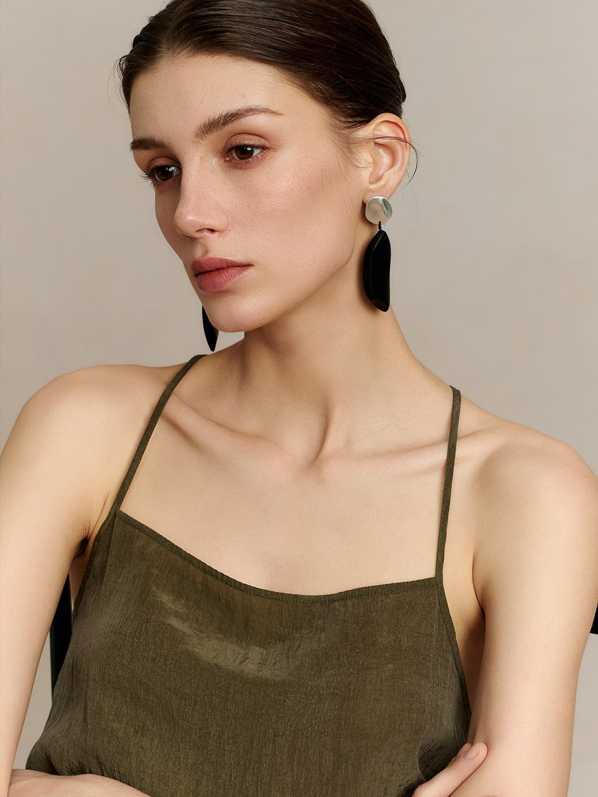 Model in a deep green thin-strap dress wearing Fairyokai Quiet Leaf silver disc and ebony leaf earrings, looking down and to the right with her hands clasped in front of her chest, against a neutral background