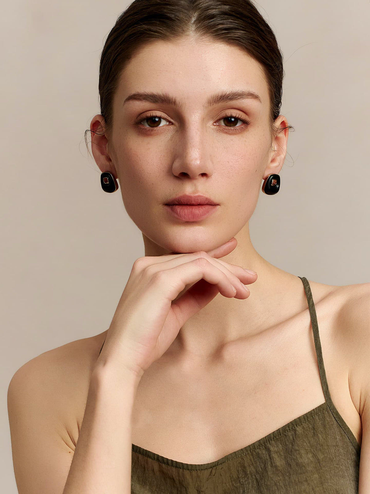 Model in a deep green thin-strap dress wearing Fairyokai Resting Corner trapezoid ebony earrings, resting her chin on her hand while looking at the camera — front view, on a neutral background