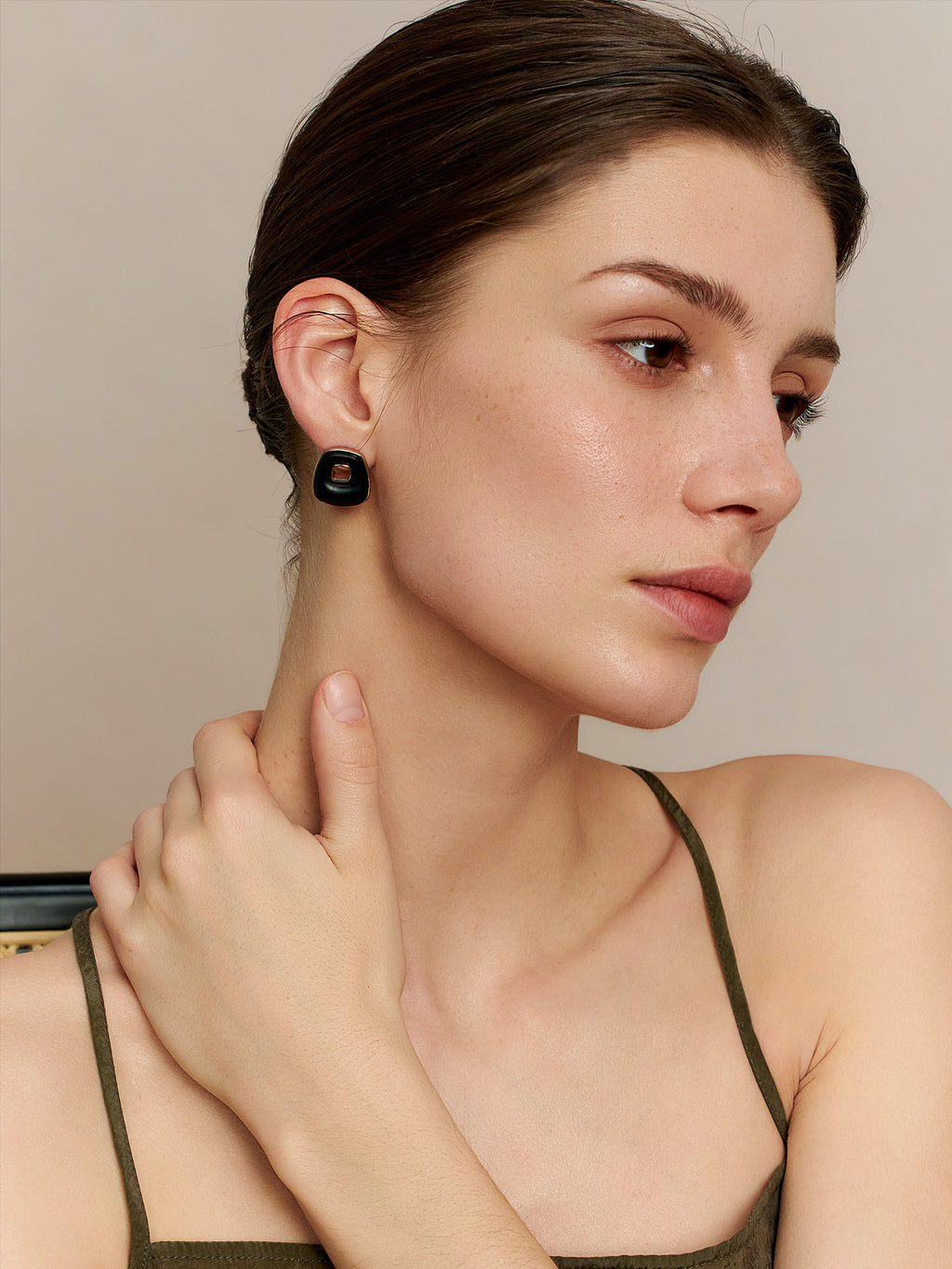 Side profile of a model in a deep green thin-strap dress, looking to the upper left, left hand touching her right neck, wearing Fairyokai Resting Corner trapezoid ebony earrings, side view on a neutral background