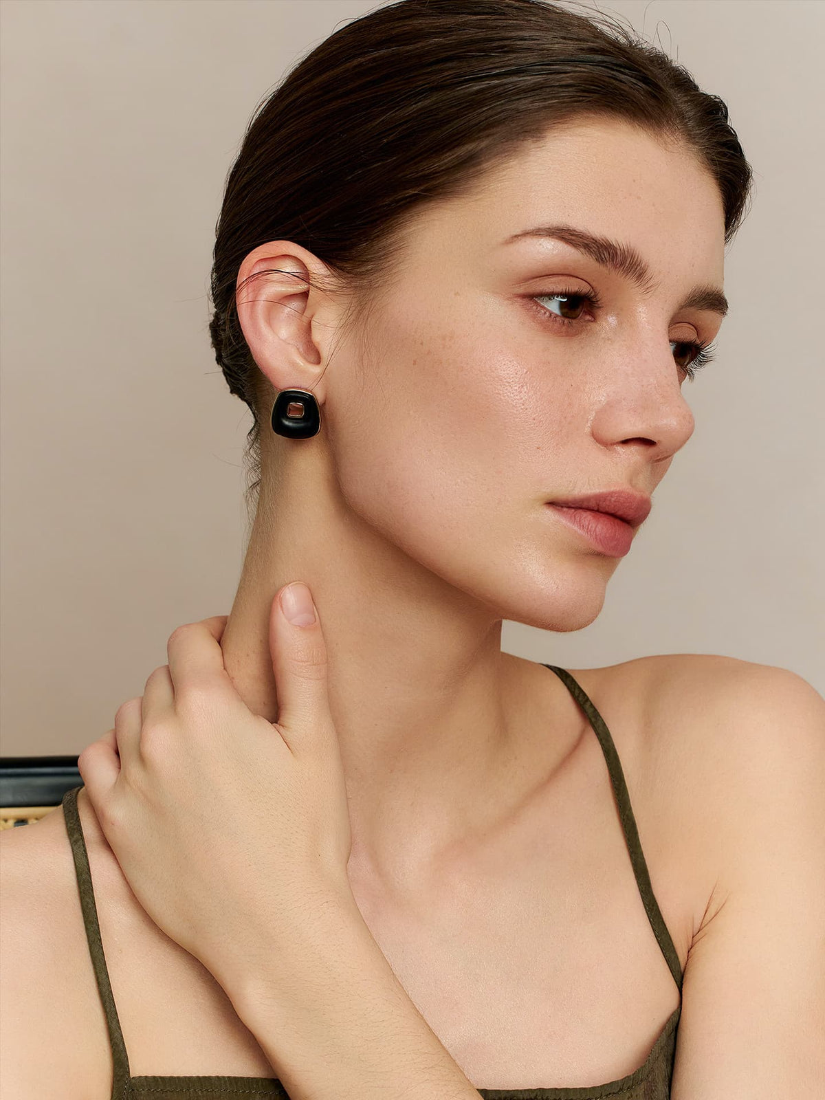 Side profile of a model in a deep green thin-strap dress, looking to the upper left, left hand touching her right neck, wearing Fairyokai Resting Corner trapezoid ebony earrings, side view on a neutral background