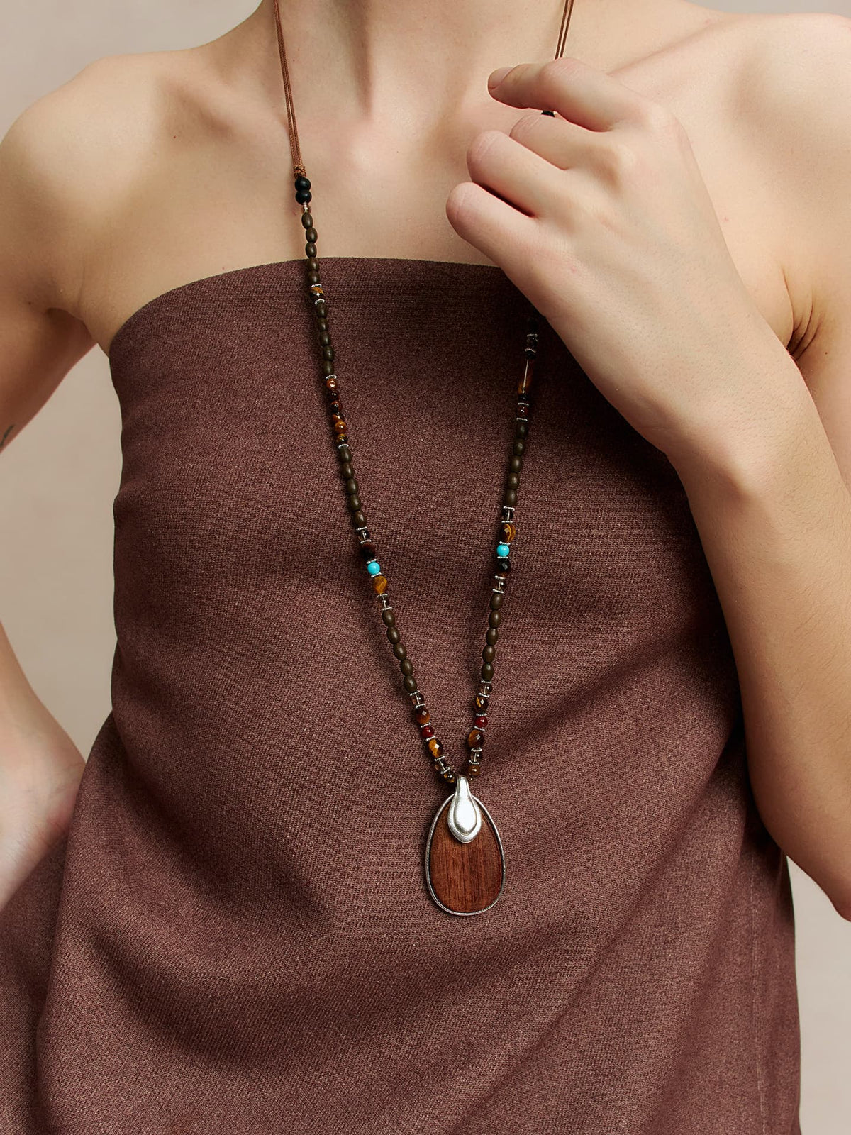 Close-up of a model wearing Fairyokai Return to Nature wood pendant necklace, brown strapless top, left hand touching the necklace, framed at chest level.