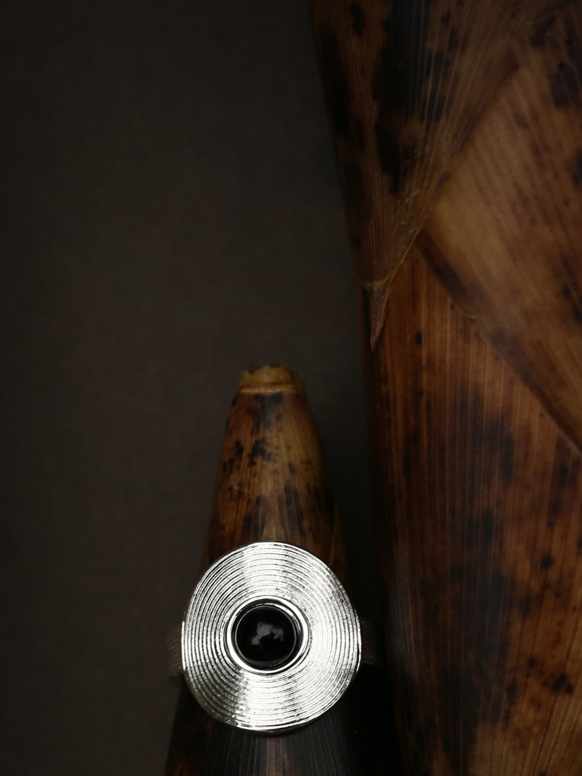 Close-up of Fairyokai Returning Depth black agate and silver band ring placed on a bamboo shoot-shaped display object, detail view, on black background.