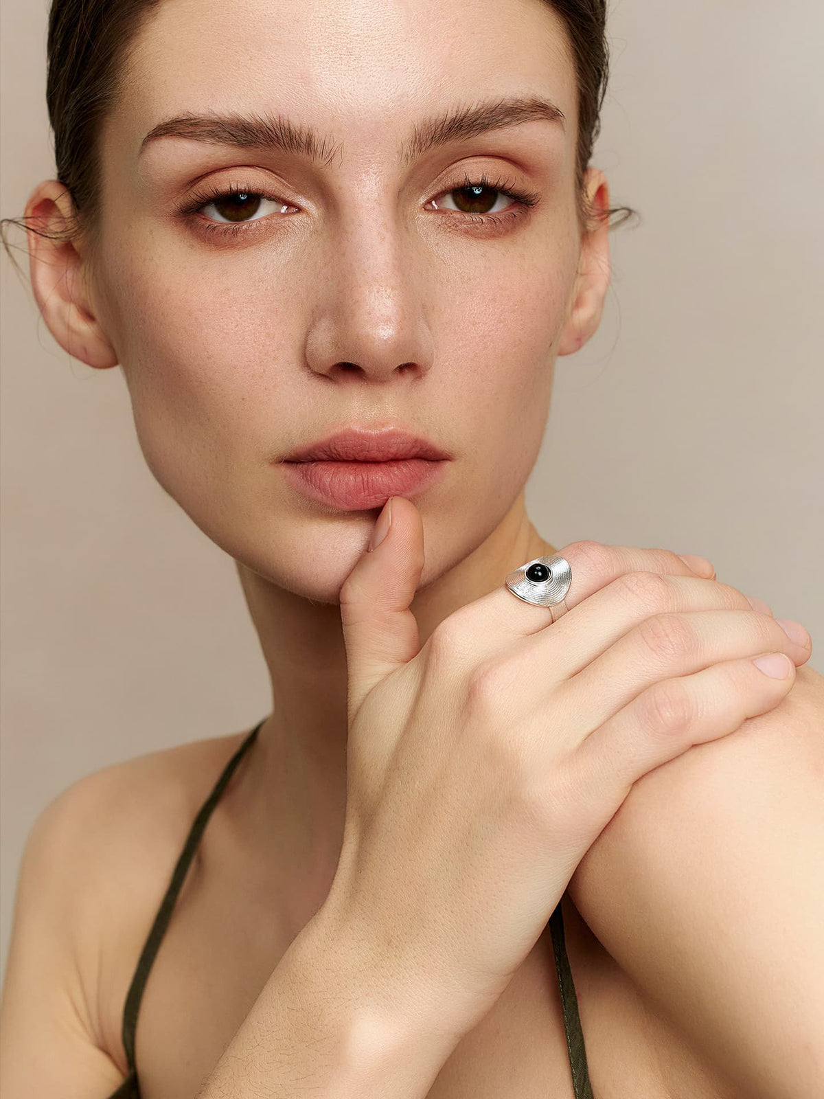 Full-length shot of a model in a thin-strap dress, wearing Fairyokai Returning Depth black agate and silver band ring, right hand resting on left shoulder with fingers touching her chin, on a beige background