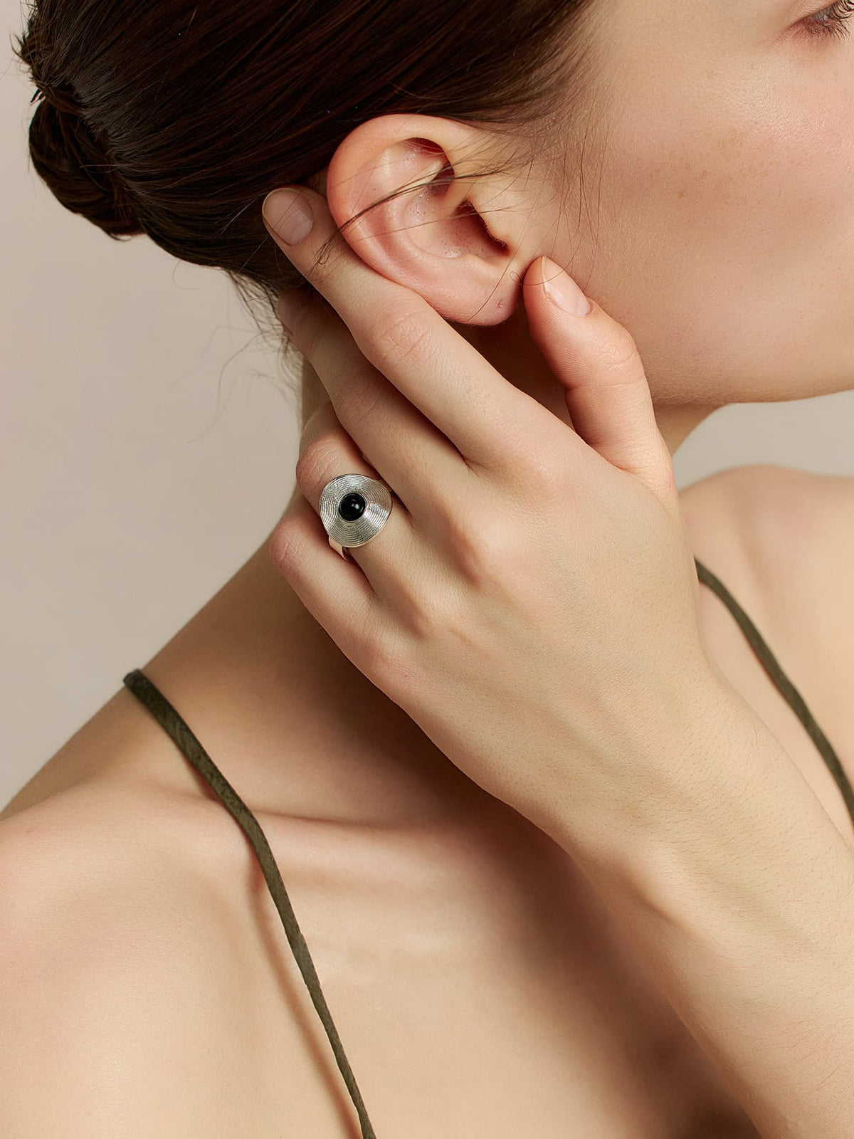 Close-up of a model in a thin-strap dress, left hand touching her right ear, head tilted to the left, focusing on the Fairyokai Returning Depth black agate and silver band ring on her left hand, a neutral background