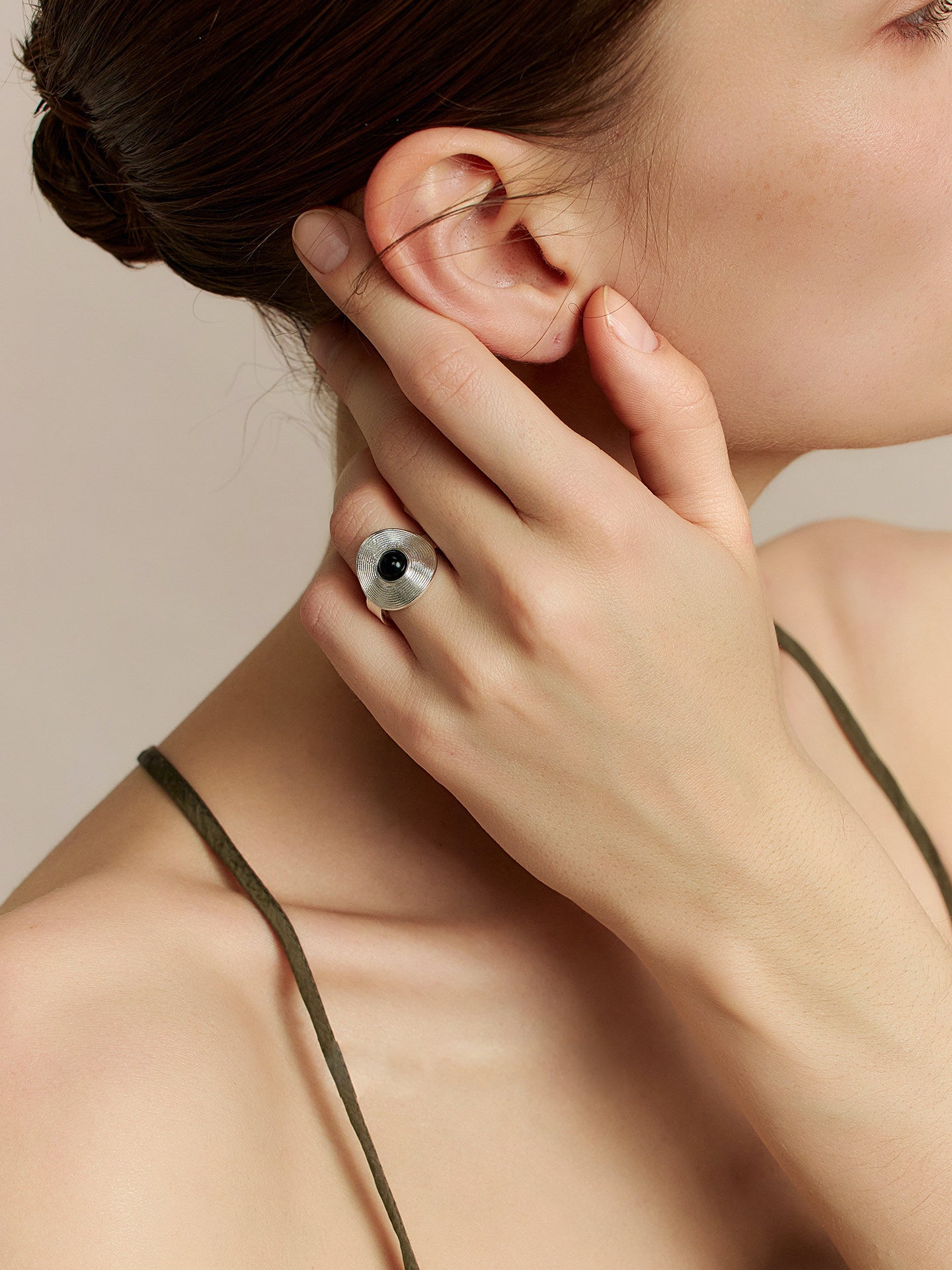 Close-up of a model in a thin-strap dress, left hand touching her right ear, head tilted to the left, focusing on the Fairyokai Returning Depth black agate and silver band ring on her left hand, a neutral background