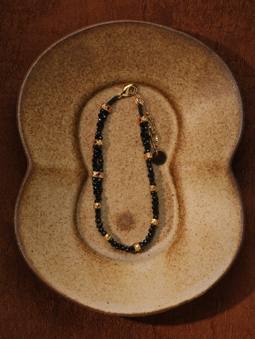 Fairyokai Rhythm Between double-layer black spinel bracelet flat-laid on a sculptural ceramic plate, showing black beads and gold-tone accents.