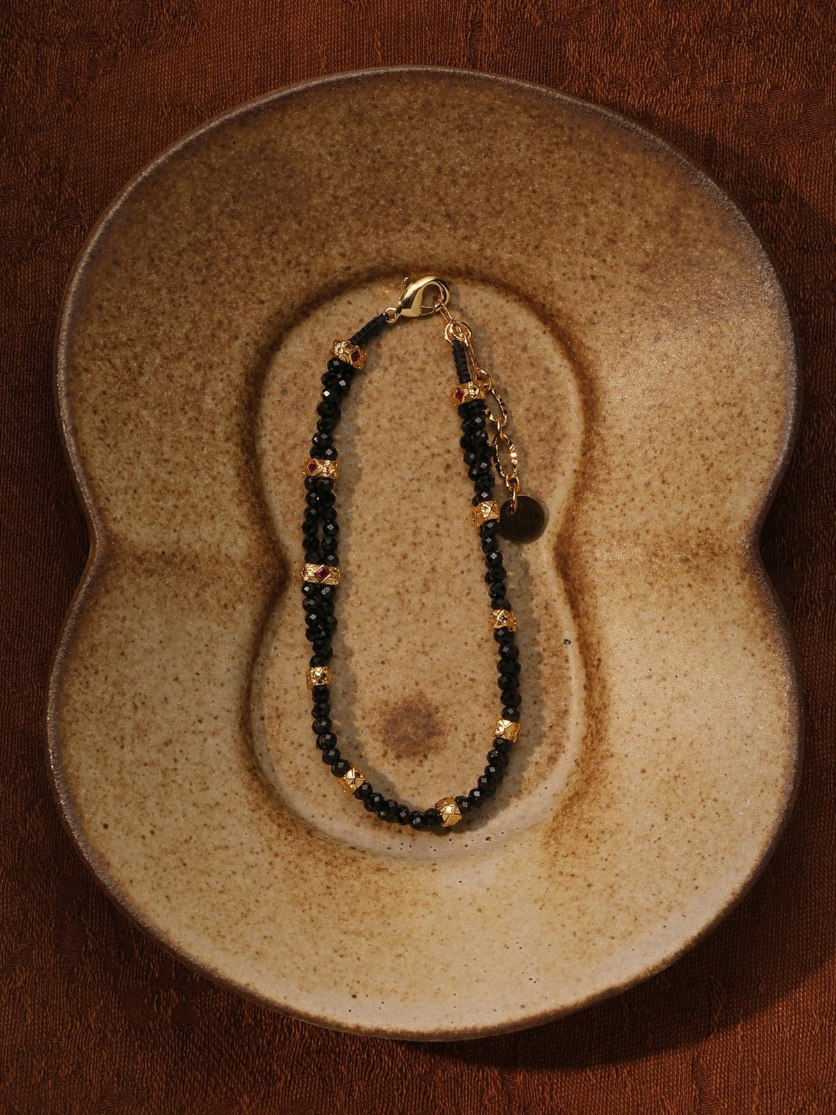 Fairyokai Rhythm Between double-layer black spinel bracelet flat-laid on a sculptural ceramic plate, showing black beads and gold-tone accents.