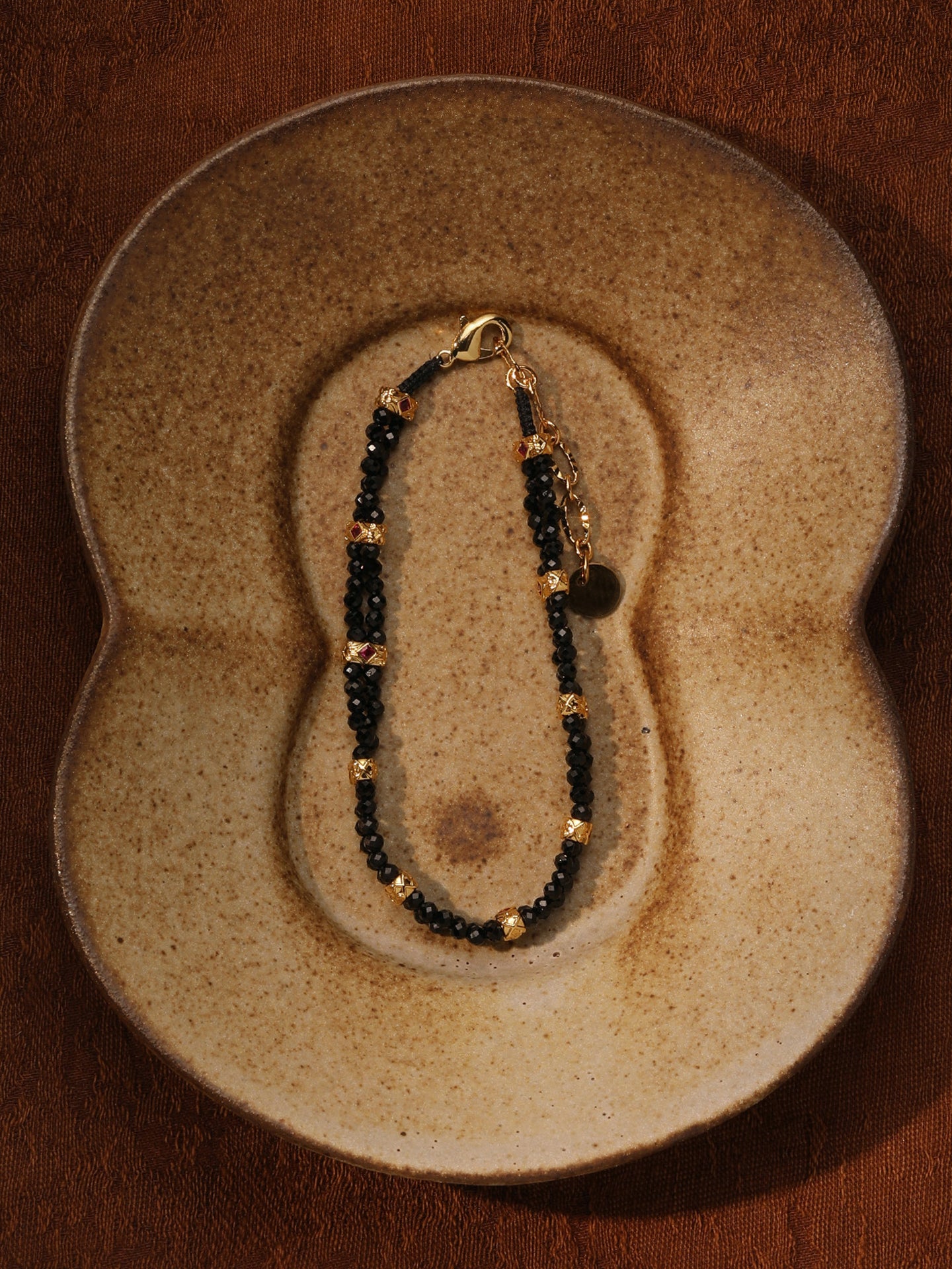 Fairyokai Rhythm Between double-layer black spinel bracelet flat-laid on a sculptural ceramic plate, showing black beads and gold-tone accents.
