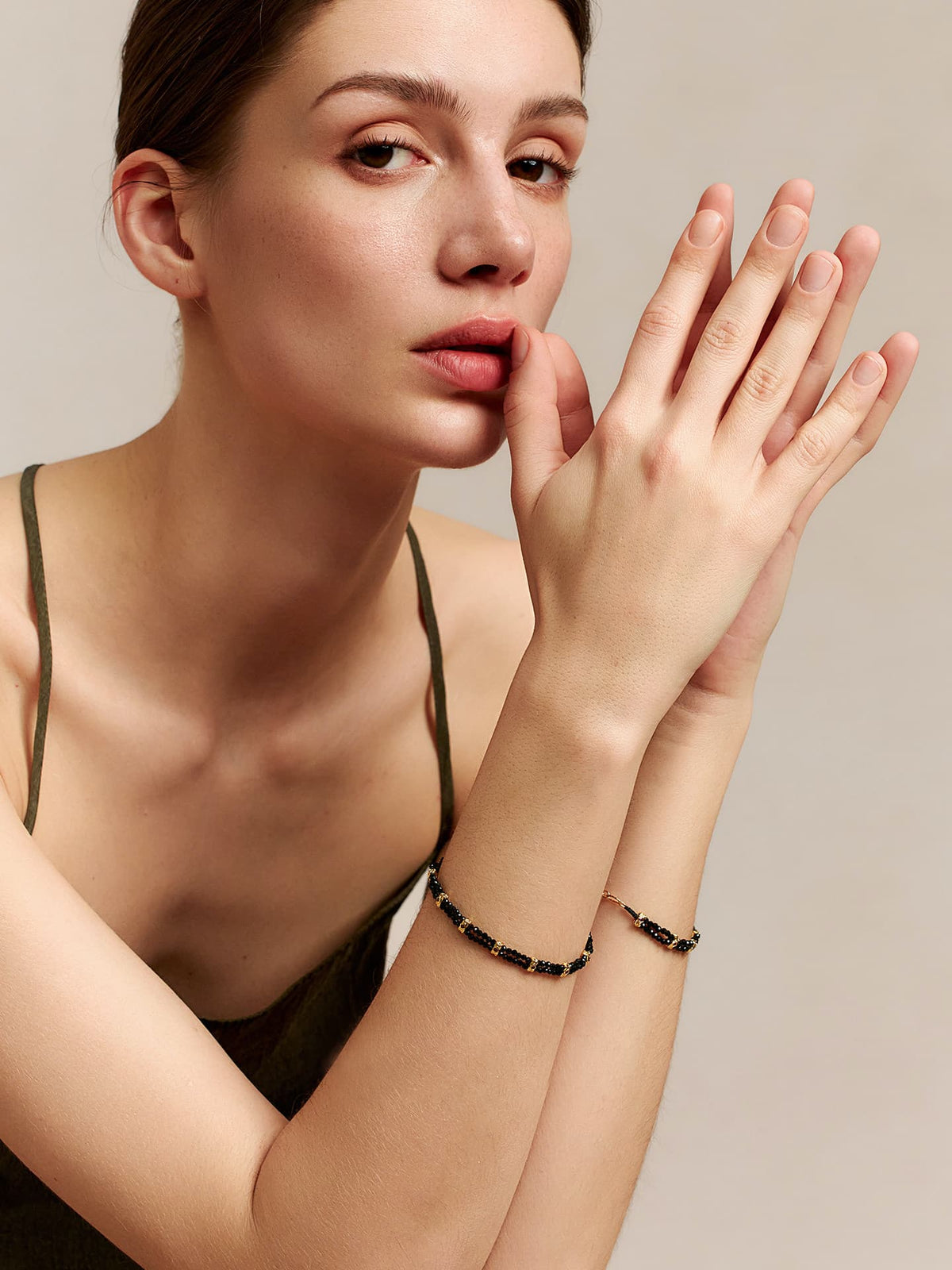 Model wearing the Fairyokai Rhythm Between black spinel bracelet on both wrists, hands raised near the face on a beige background.