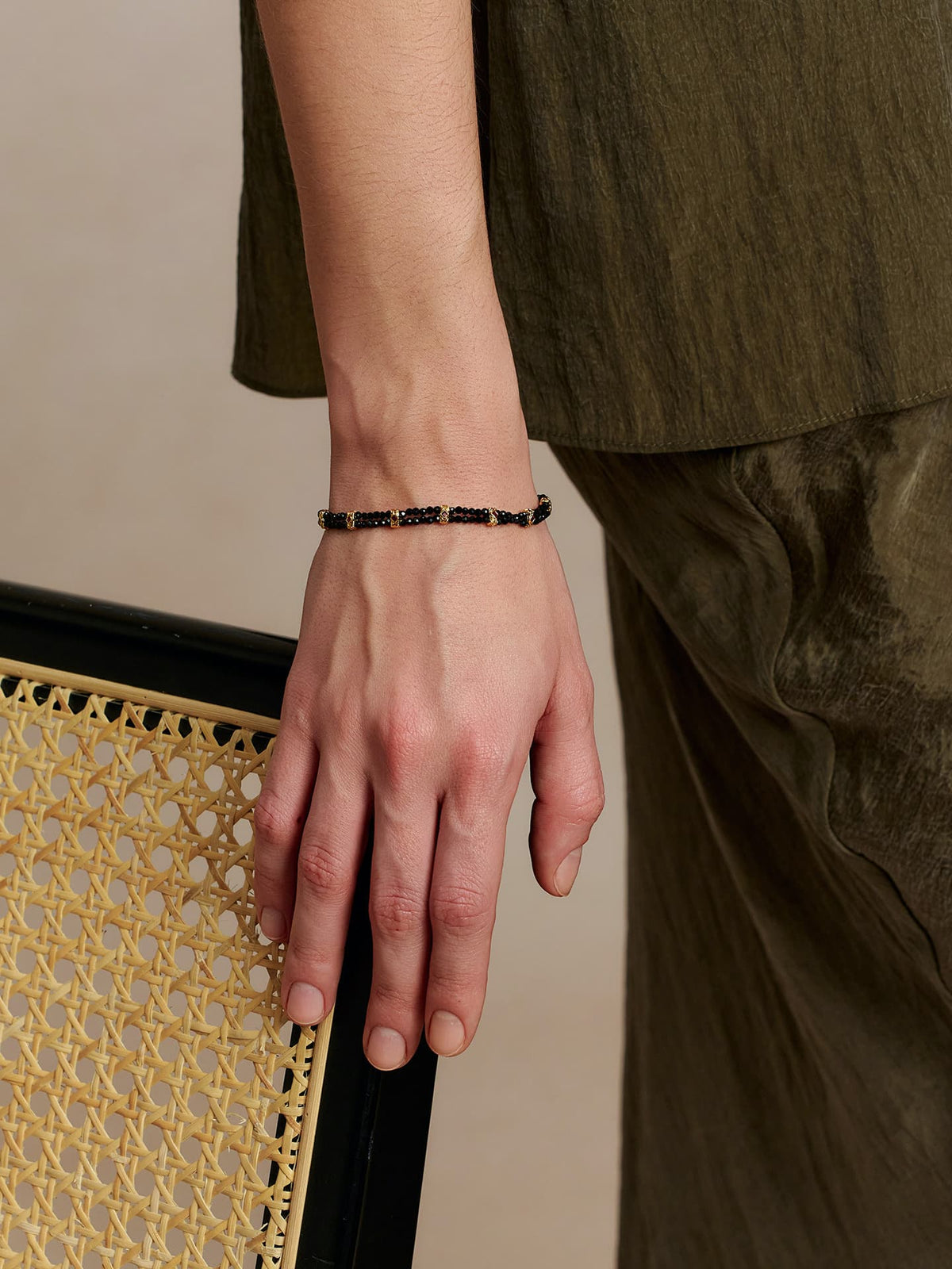Close-up of the Fairyokai Rhythm Between black spinel bracelet worn on the wrist beside a woven chair and olive fabric.