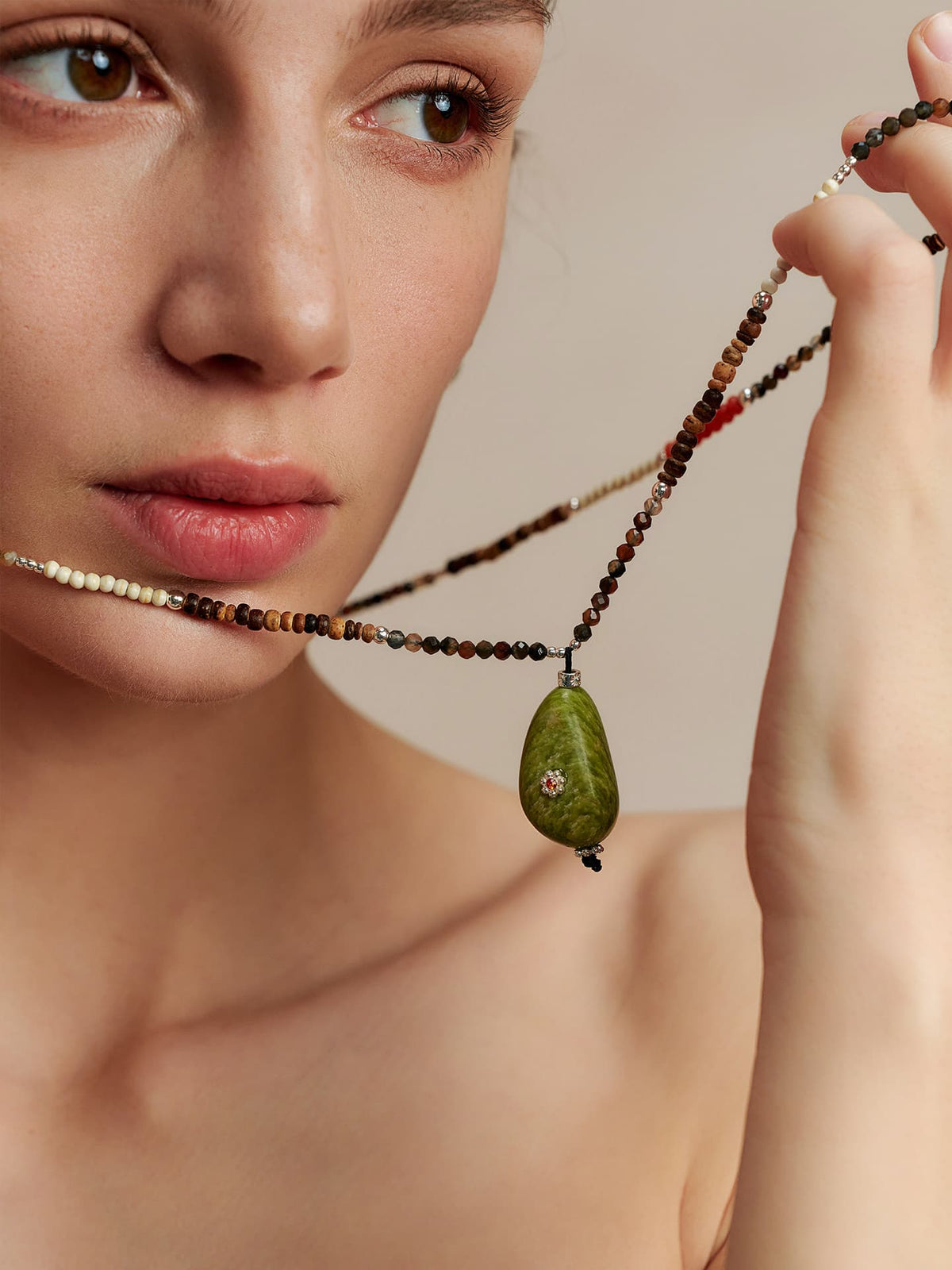 Model holding the Fairyokai Spring Sequence multicolor avocado necklace with fingers raised, looking to the upper left, close-up from collarbone up, fully showing the necklace.